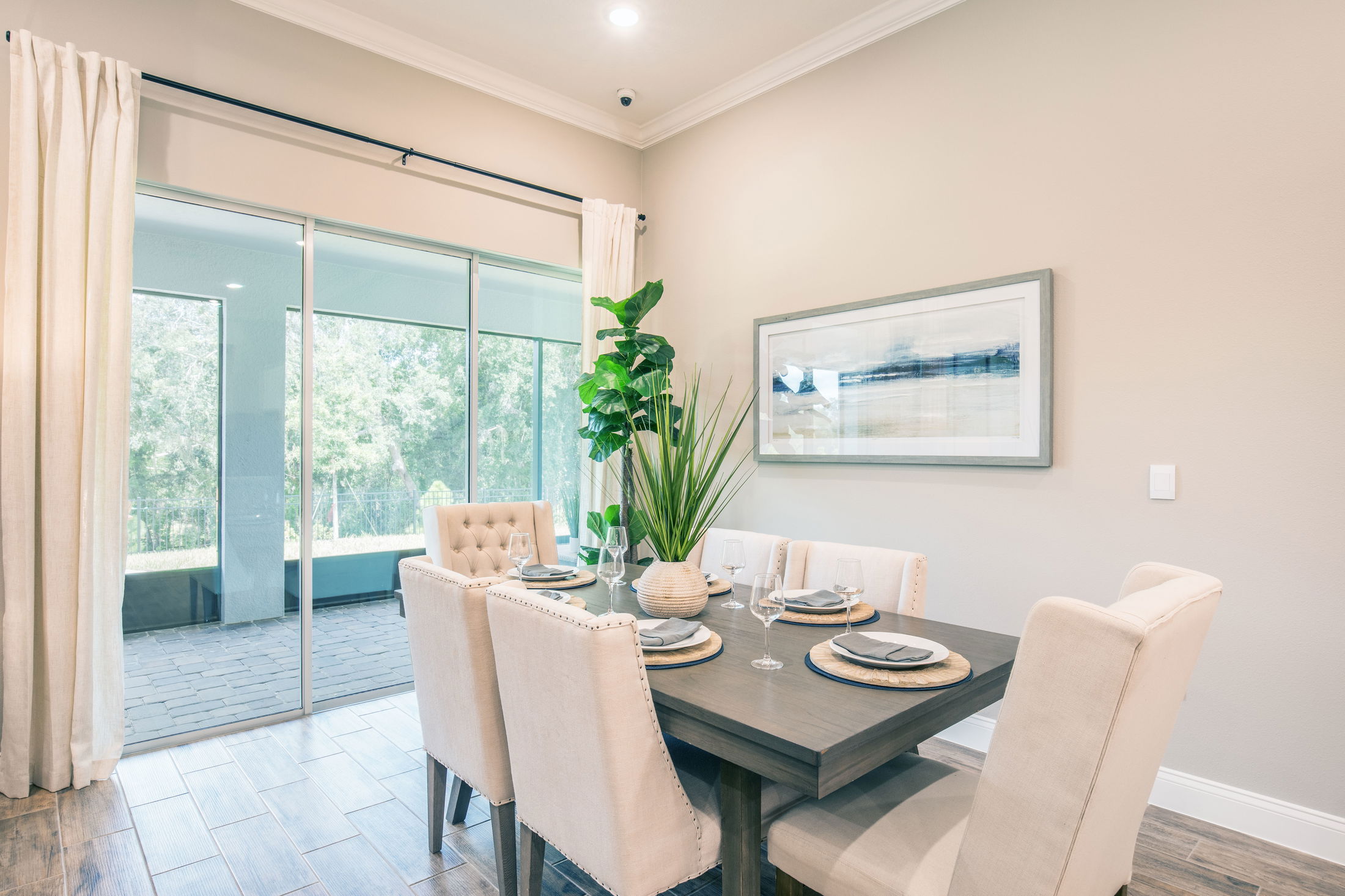 Elegant dining room with a modern table setting, cream chairs, and a large potted plant, adjacent to a glass sliding door with a scenic outdoor view.