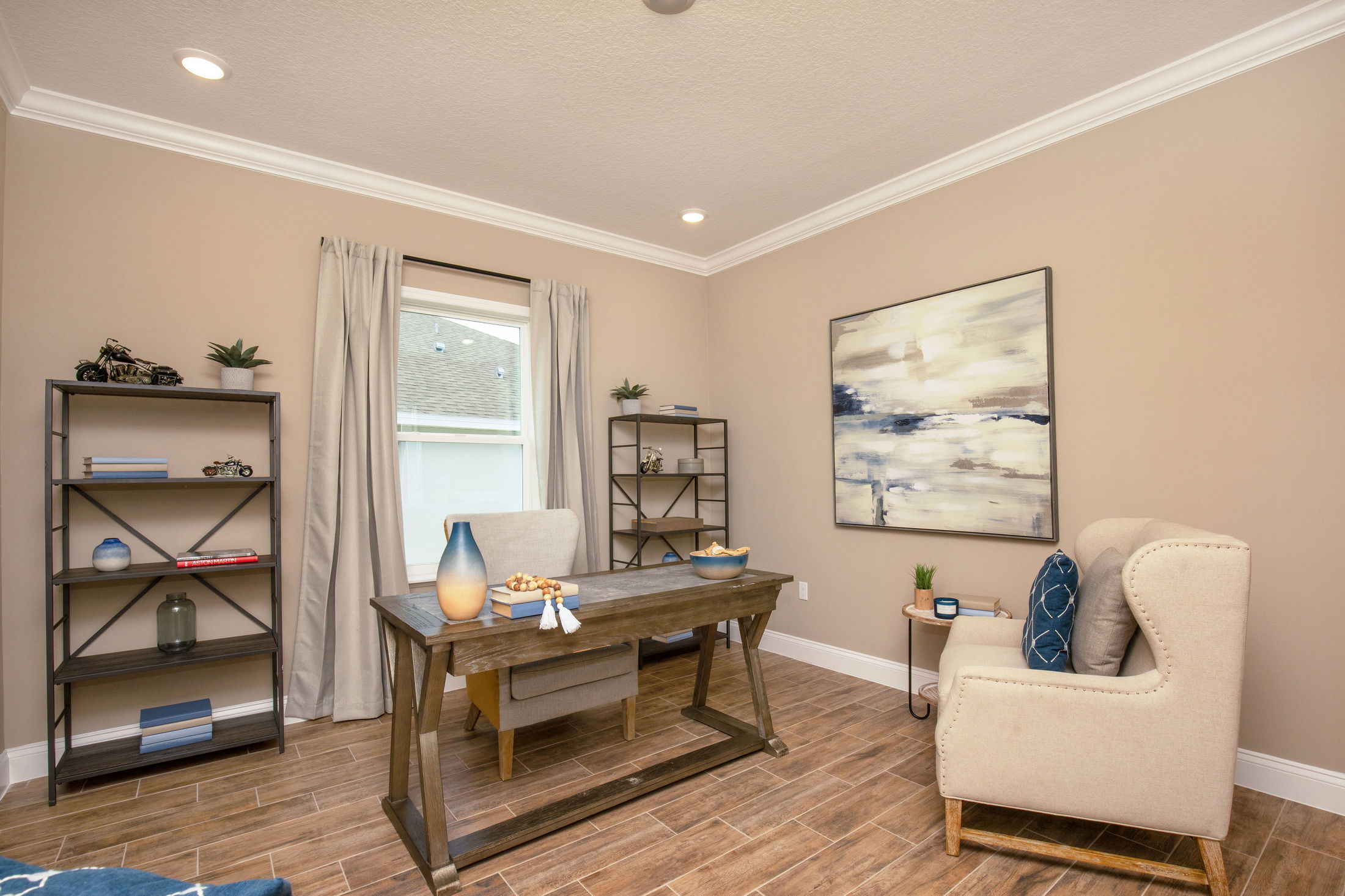 Modern home office with a wooden desk, beige armchair, bookshelves, abstract wall art, and natural lighting.