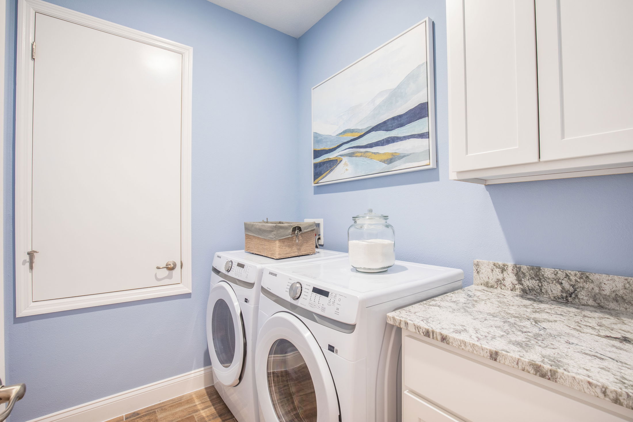 Modern laundry room with blue walls, featuring a front-loading washer and dryer set, white cabinets, granite countertop, and abstract wall art.