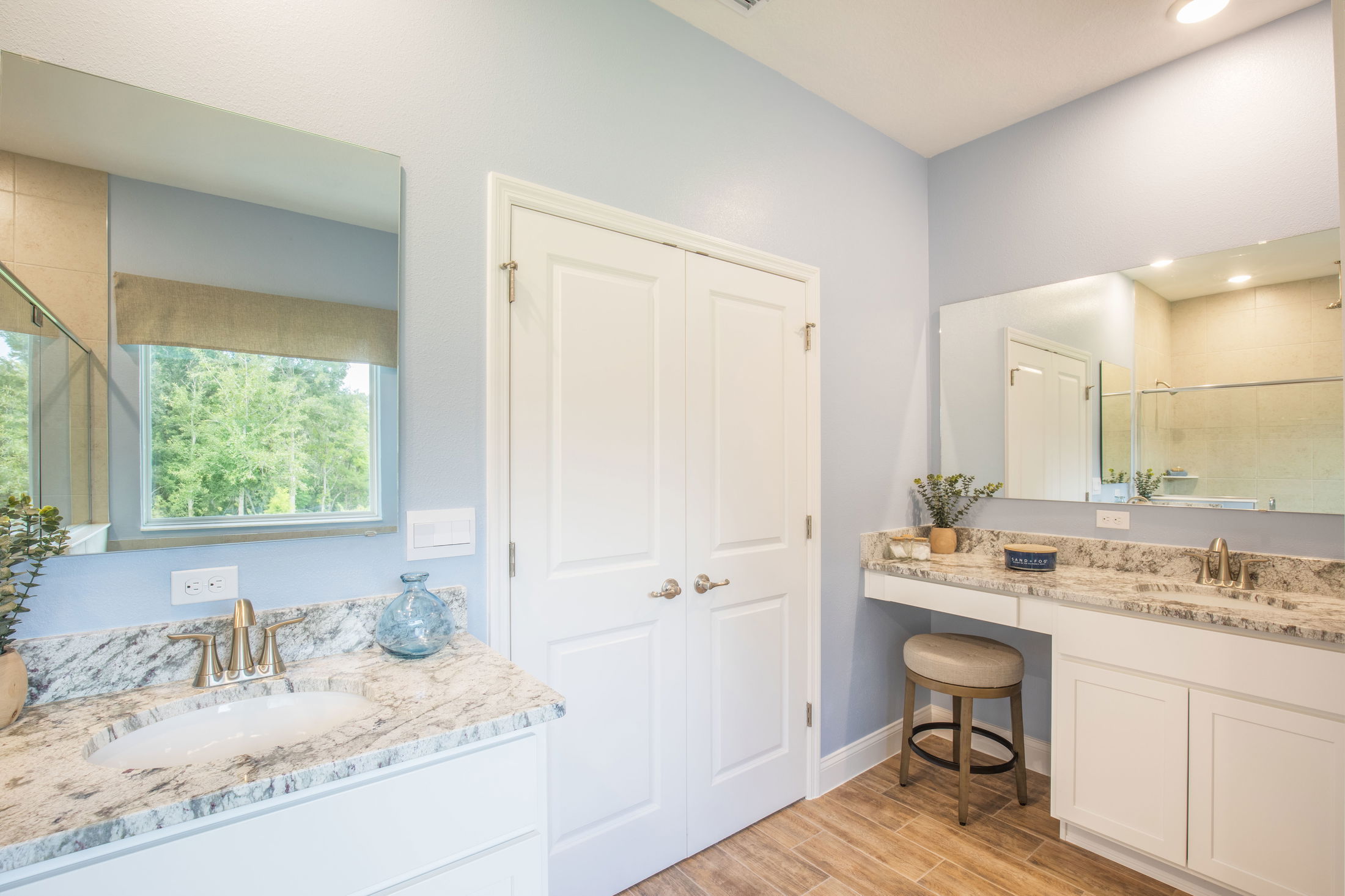Modern bathroom with marble countertops, wooden flooring, and large mirrors reflecting natural light.