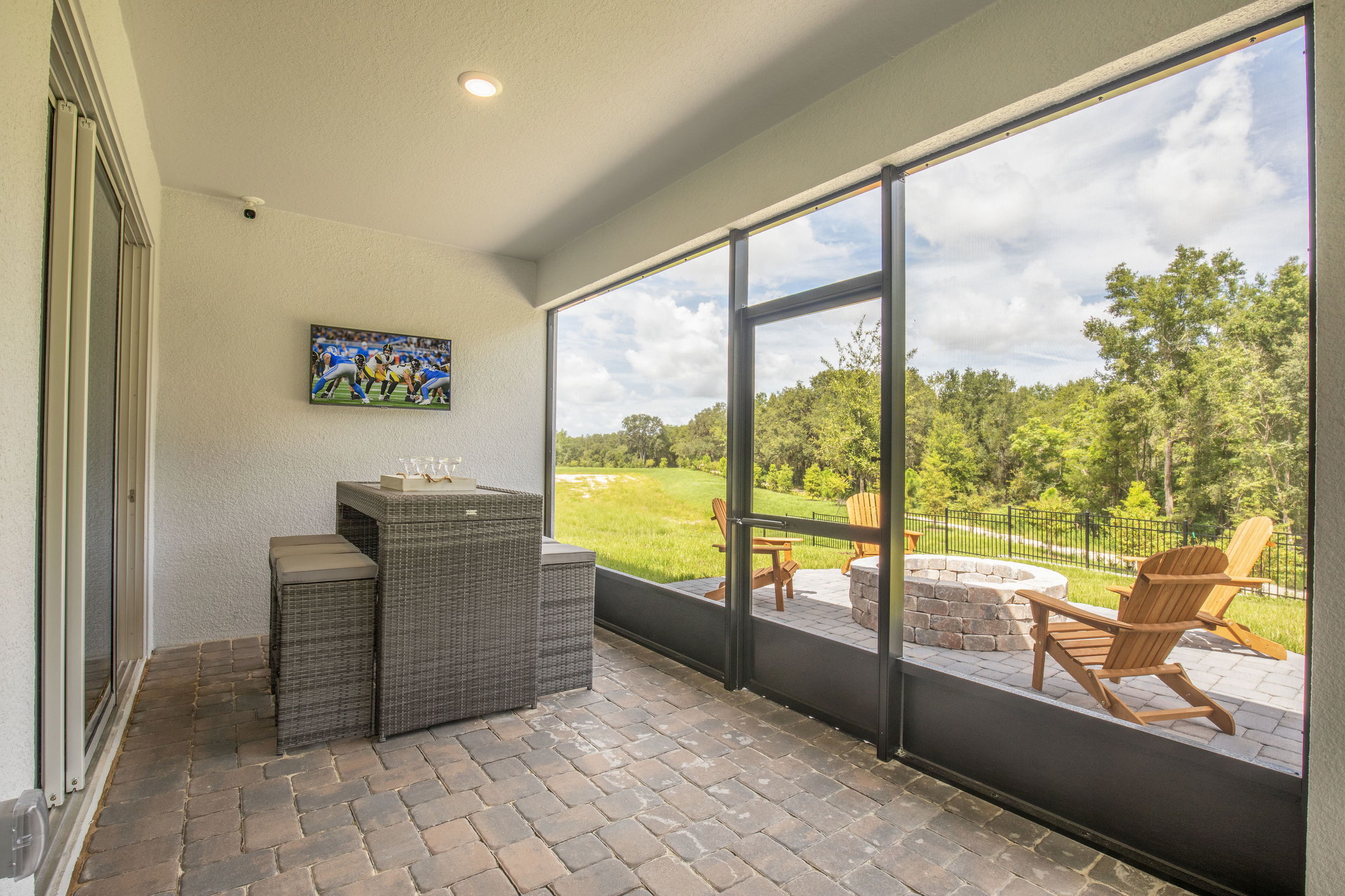 Screened-in patio with wicker furniture, wall-mounted TV, and view of a backyard with Adirondack chairs surrounding a stone fire pit.