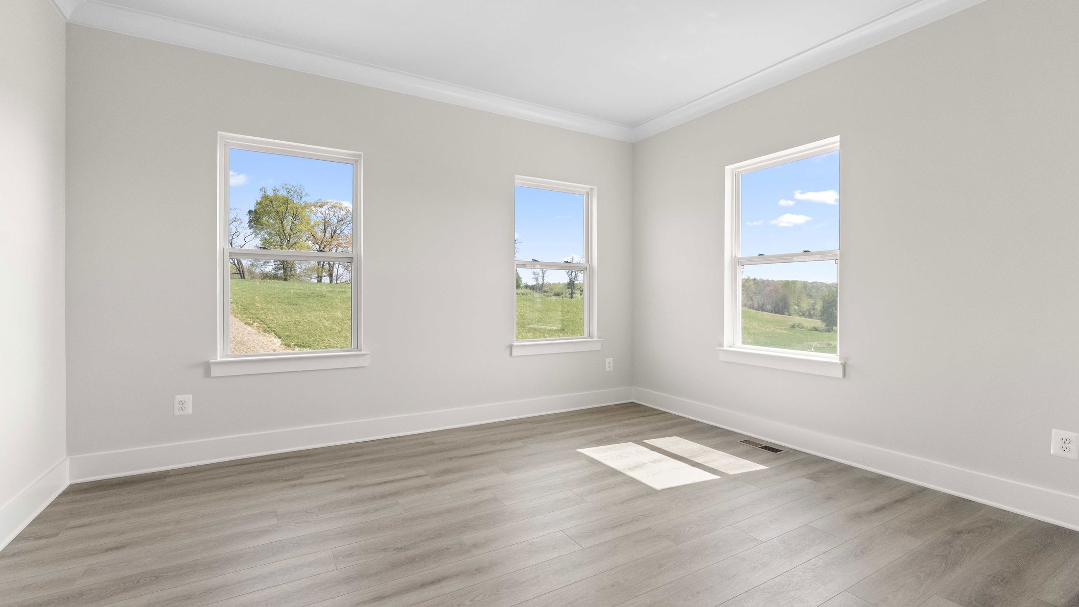 Bright, empty room with three large windows and hardwood flooring overlooking a grassy landscape.