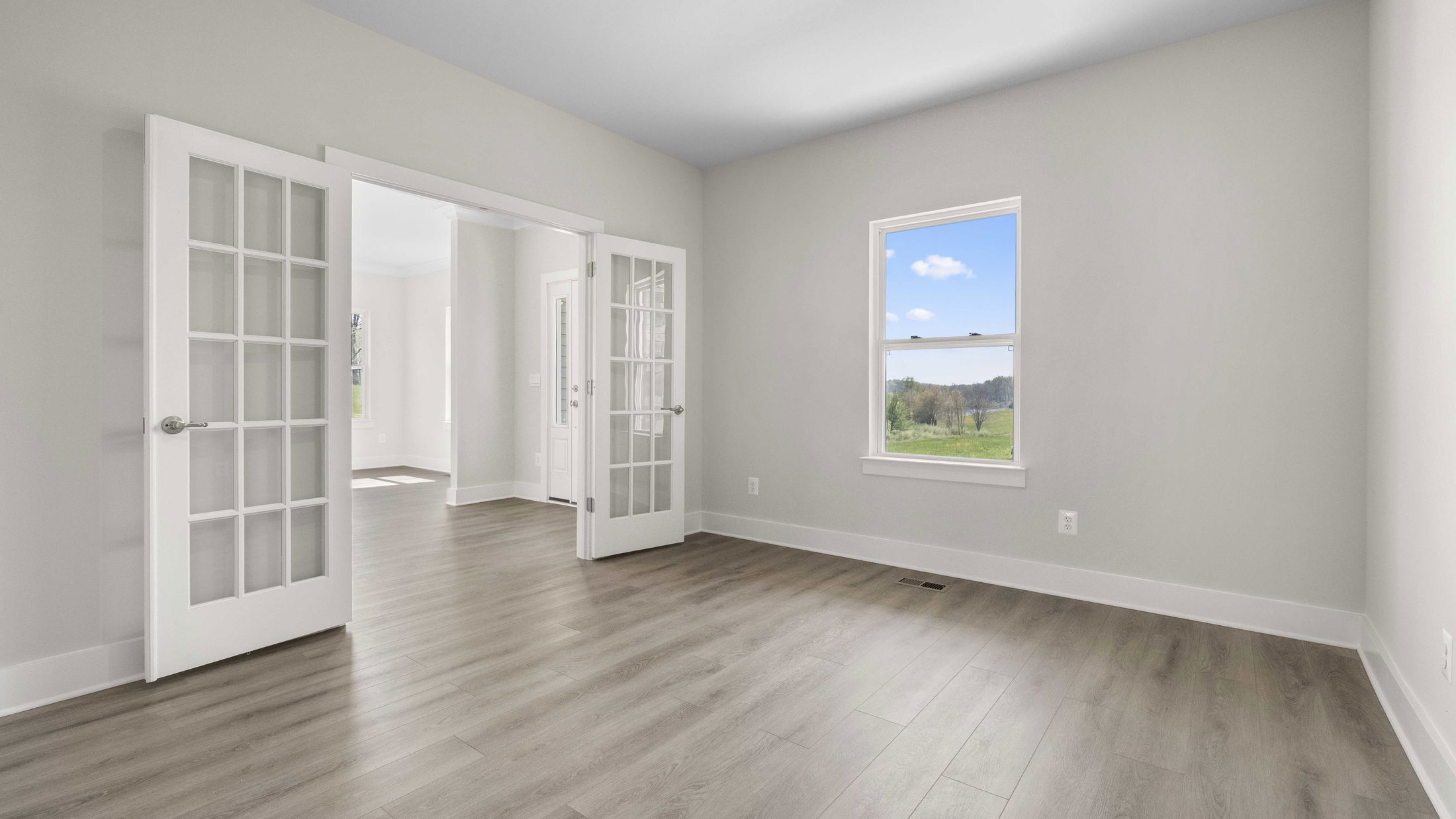 Modern empty room with light wood flooring, white walls, large window with scenic view, and French doors opening to another bright space.
