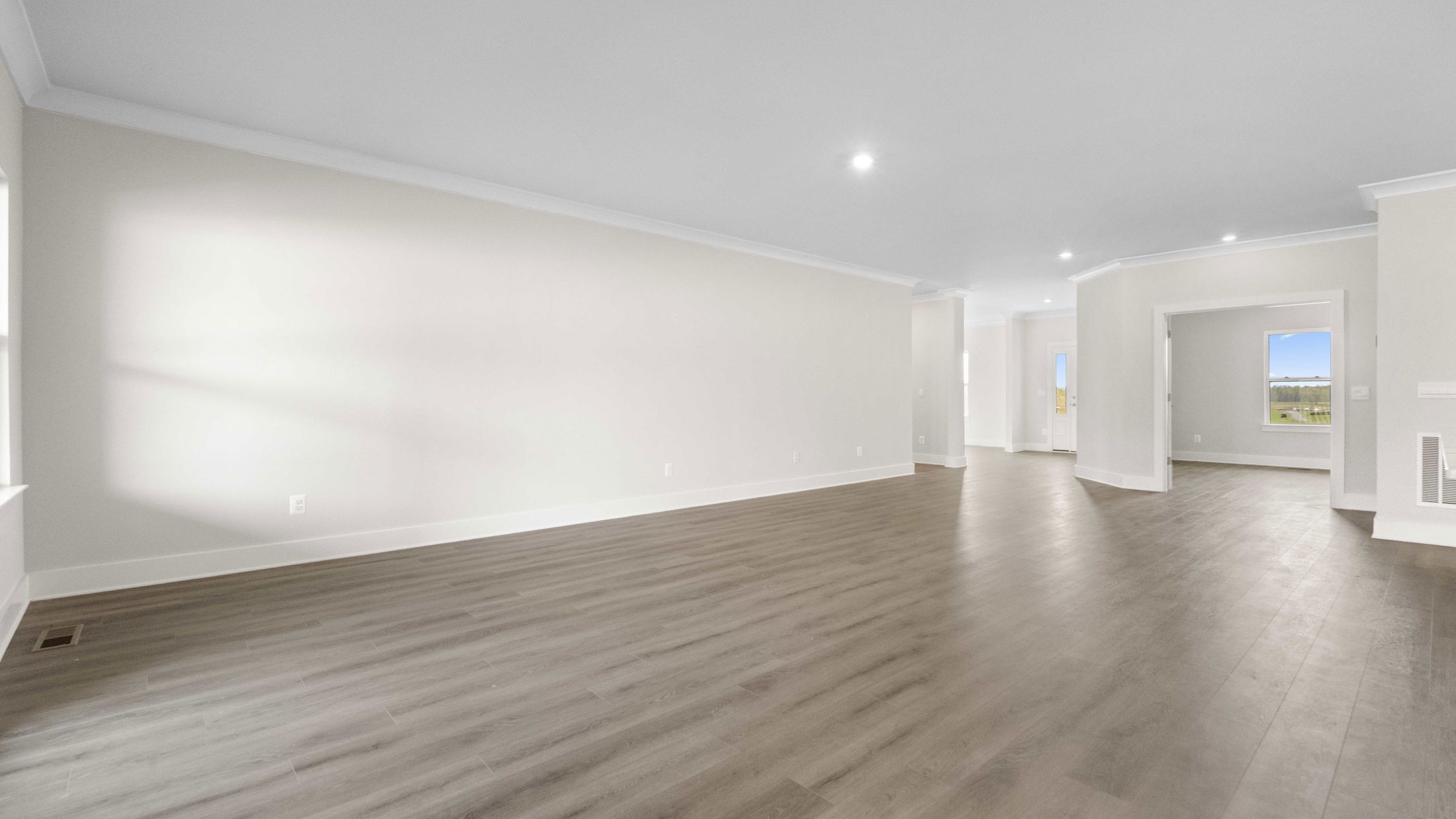 Spacious empty room with light gray walls, hardwood flooring, and natural light streaming through a window.