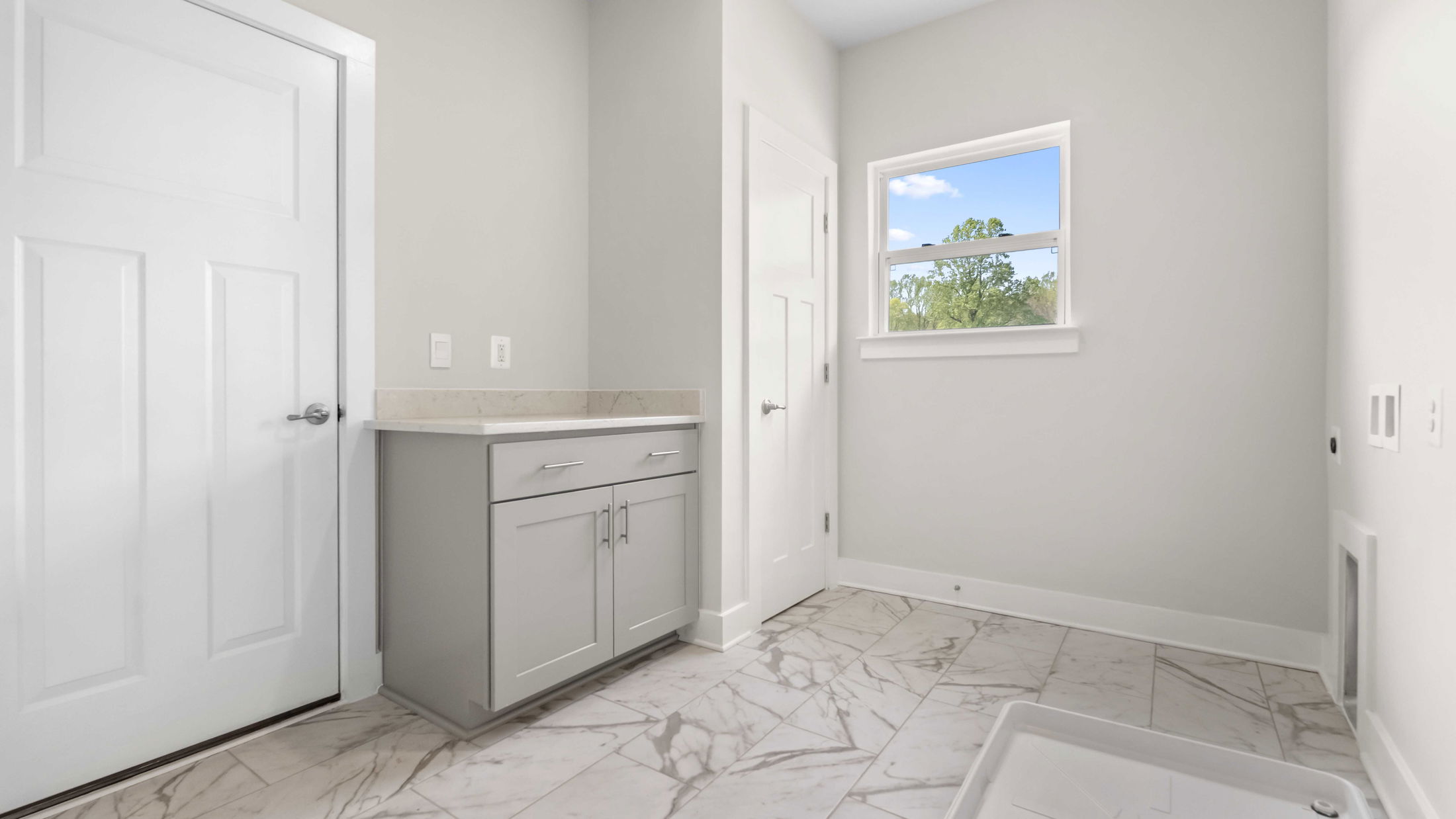 Laundry room with gray cabinets, white walls, and marble tile flooring, featuring a window and a utility sink area.