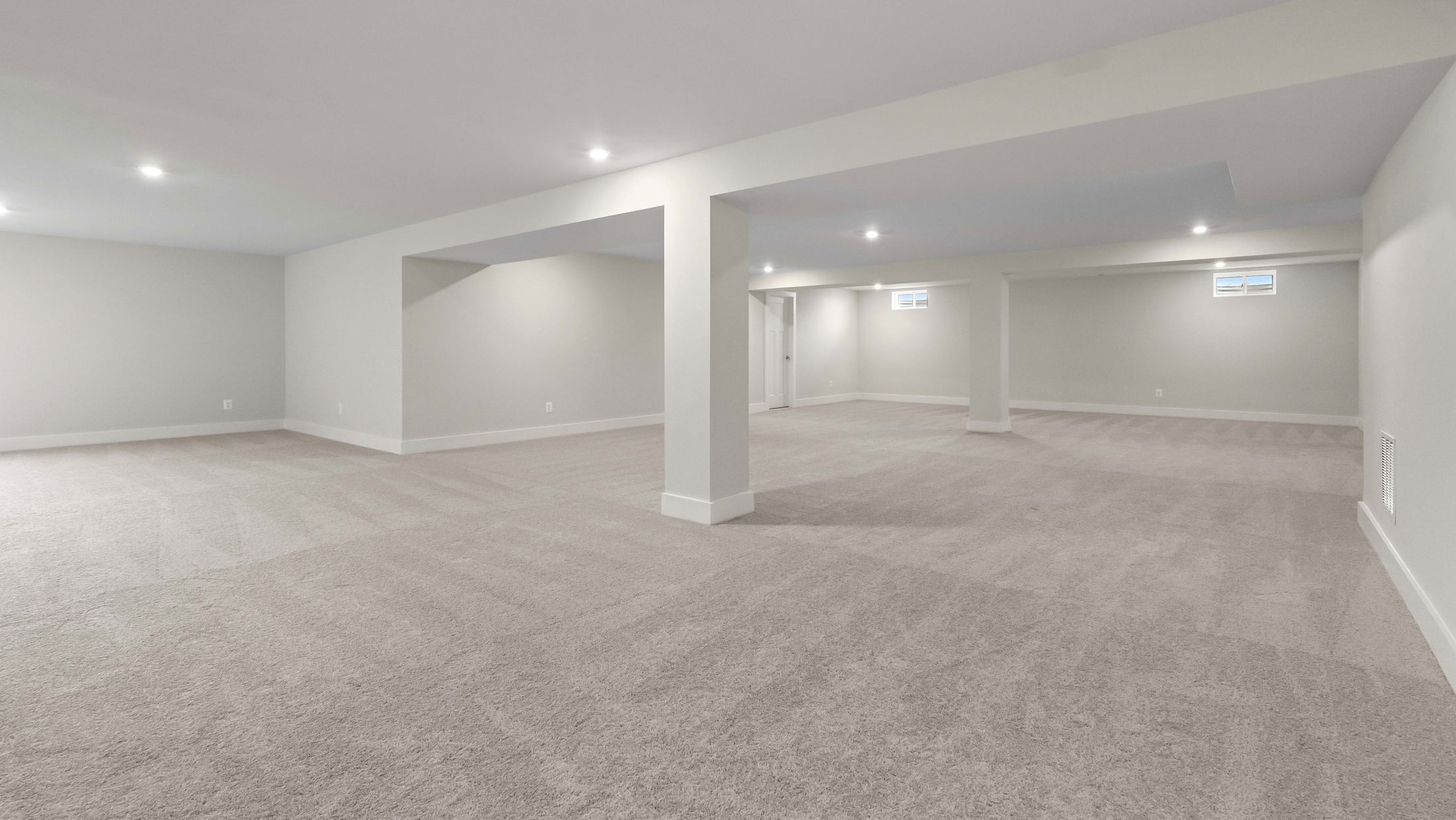 Spacious, empty basement room with soft beige carpet, white walls, and recessed ceiling lights.