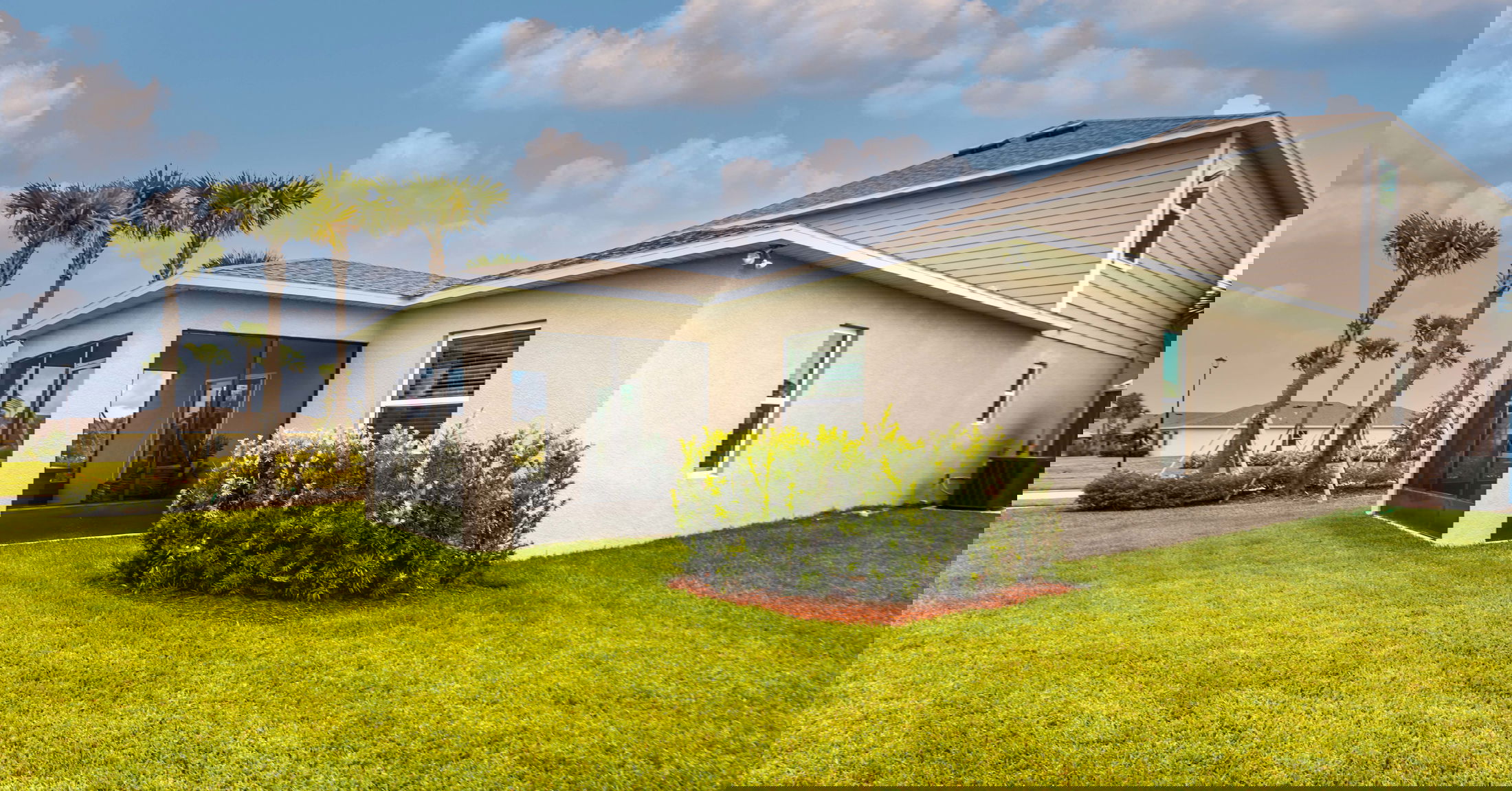 A modern house with a slanted structure surrounded by lush greenery and palm trees under a blue sky.