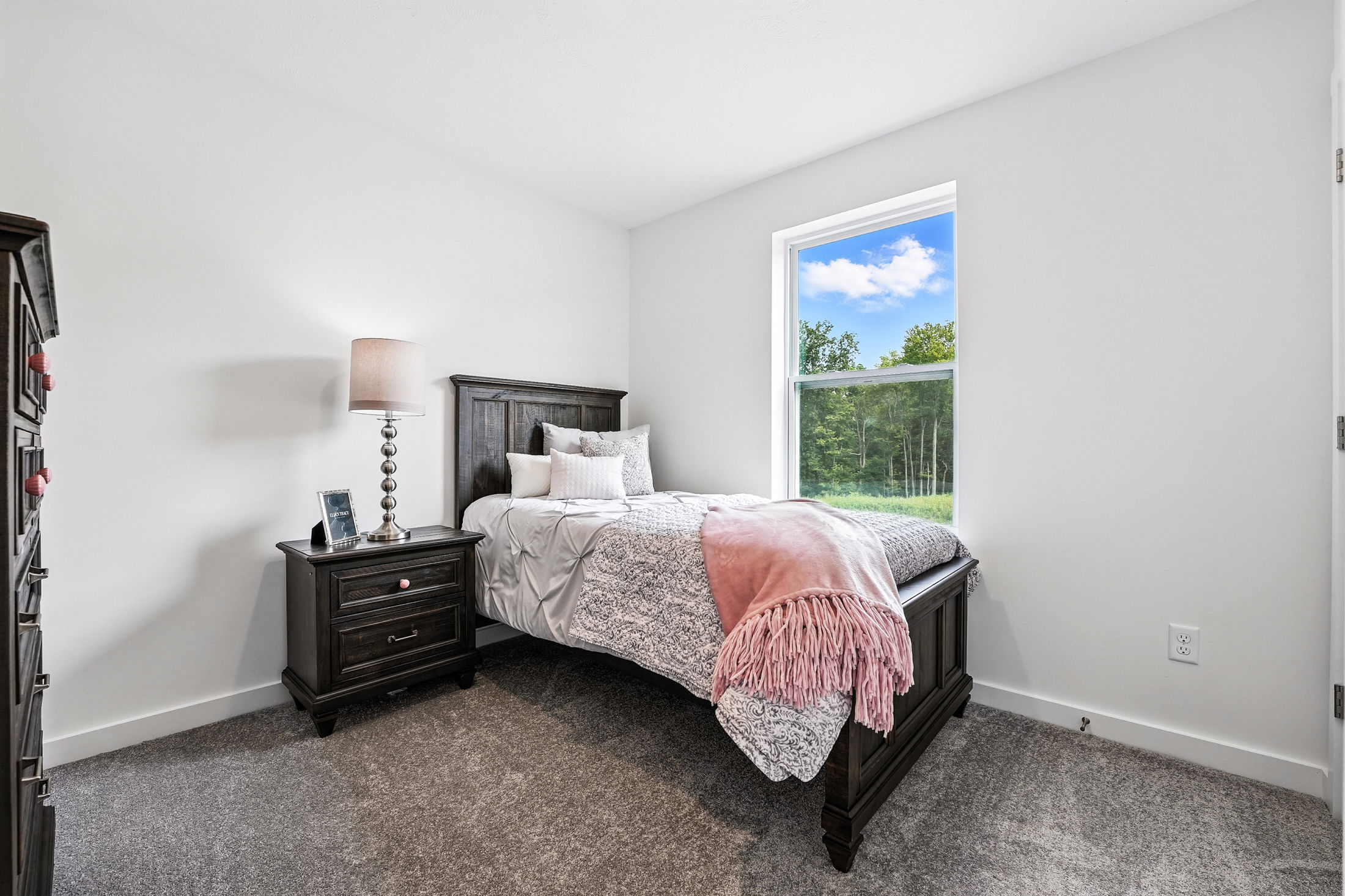 Cozy bedroom with dark wood furniture, a neatly made bed, pink throw, and a large window with a view of greenery.