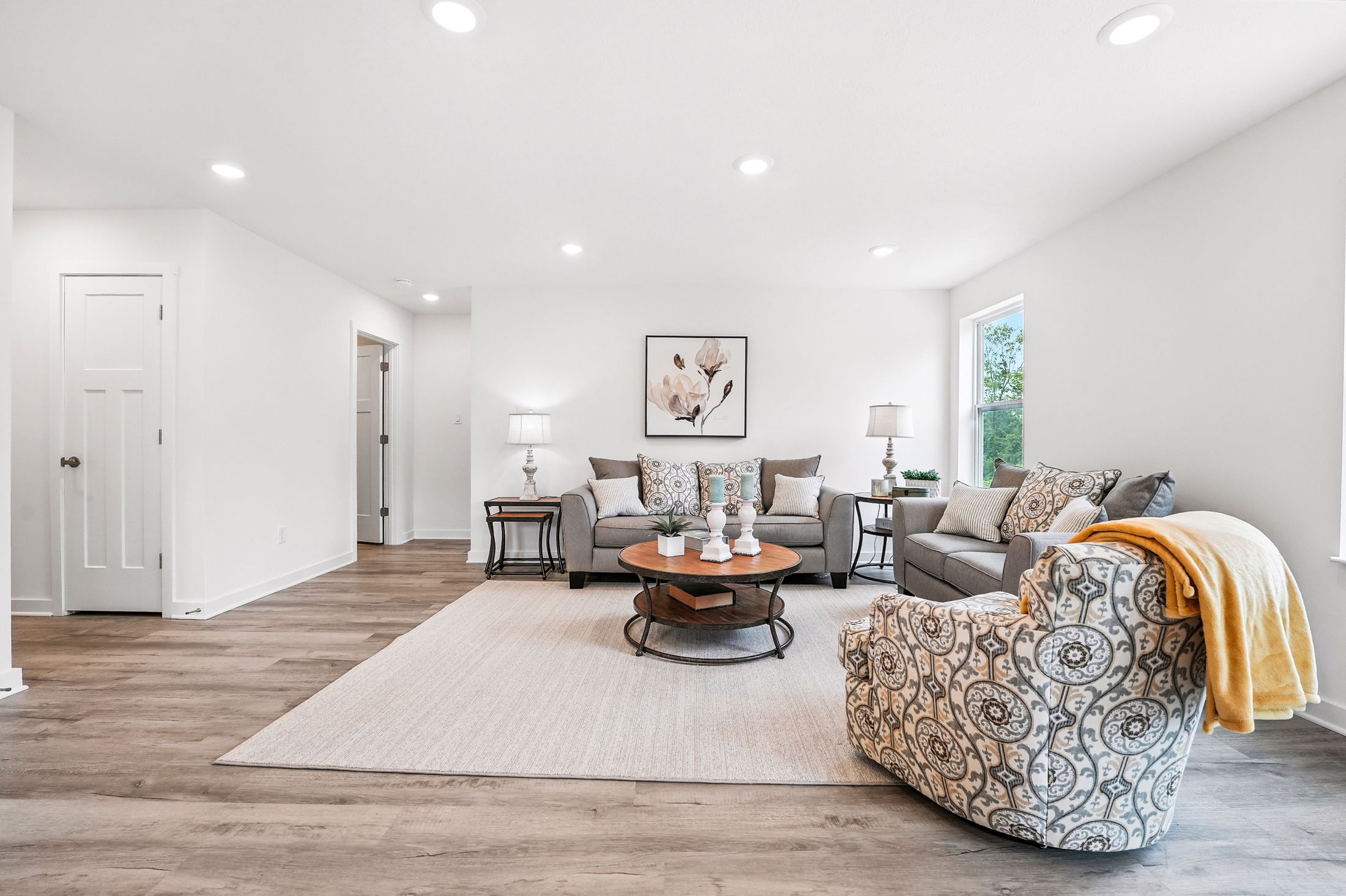 A modern living room with elegant gray sofas, a patterned armchair, and wooden coffee table, featuring neutral decor and ample natural light.