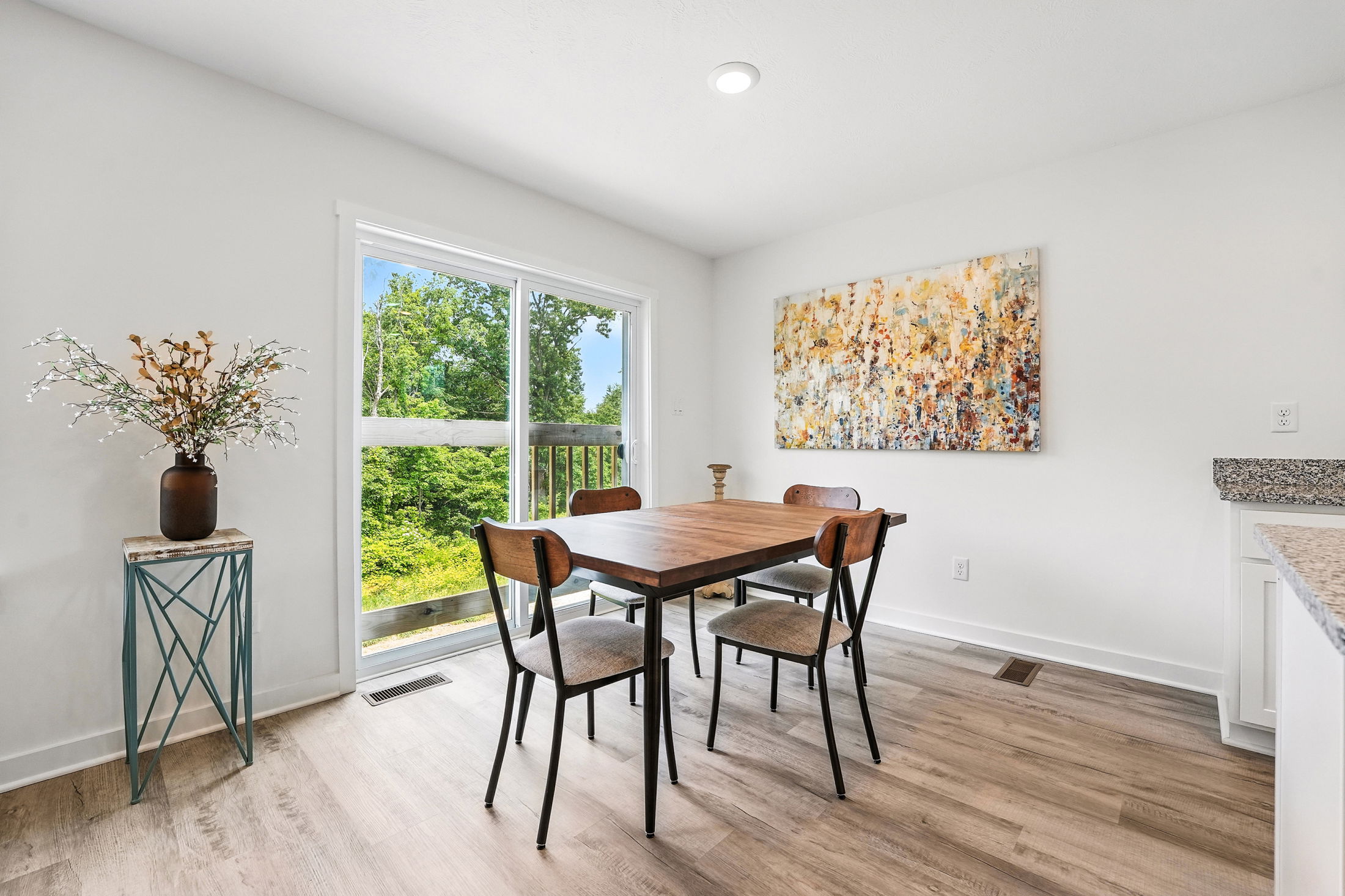 Modern dining room interior with a wooden table, four chairs, abstract wall art, and a large window overlooking lush greenery.