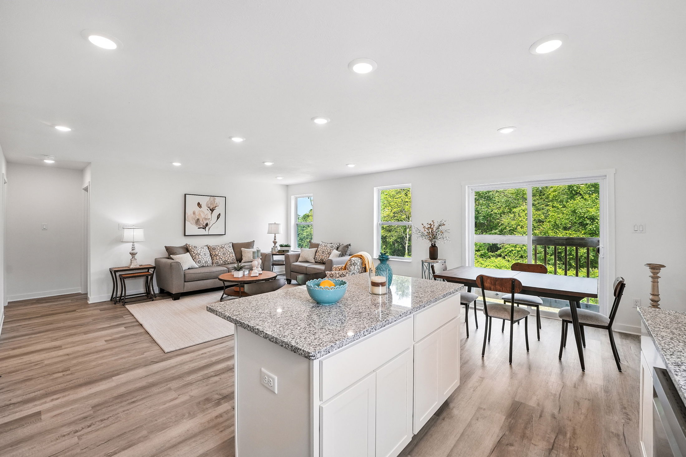 Modern living room and kitchen with open layout, featuring granite countertops, wooden flooring, stylish furniture, and large windows with a view of lush greenery.