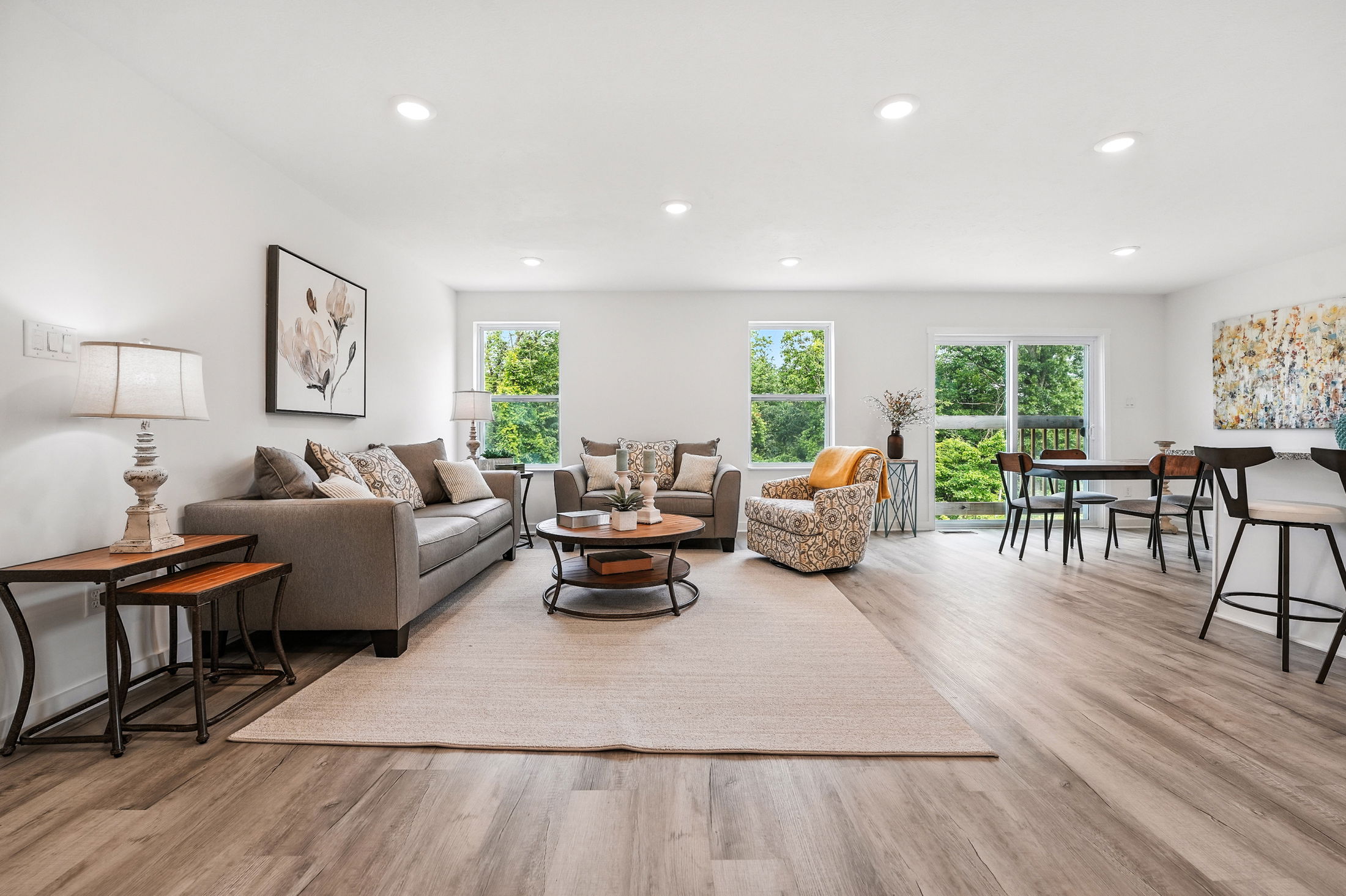 Modern living room with gray sofas, patterned armchair, wood flooring, and large windows overlooking greenery.