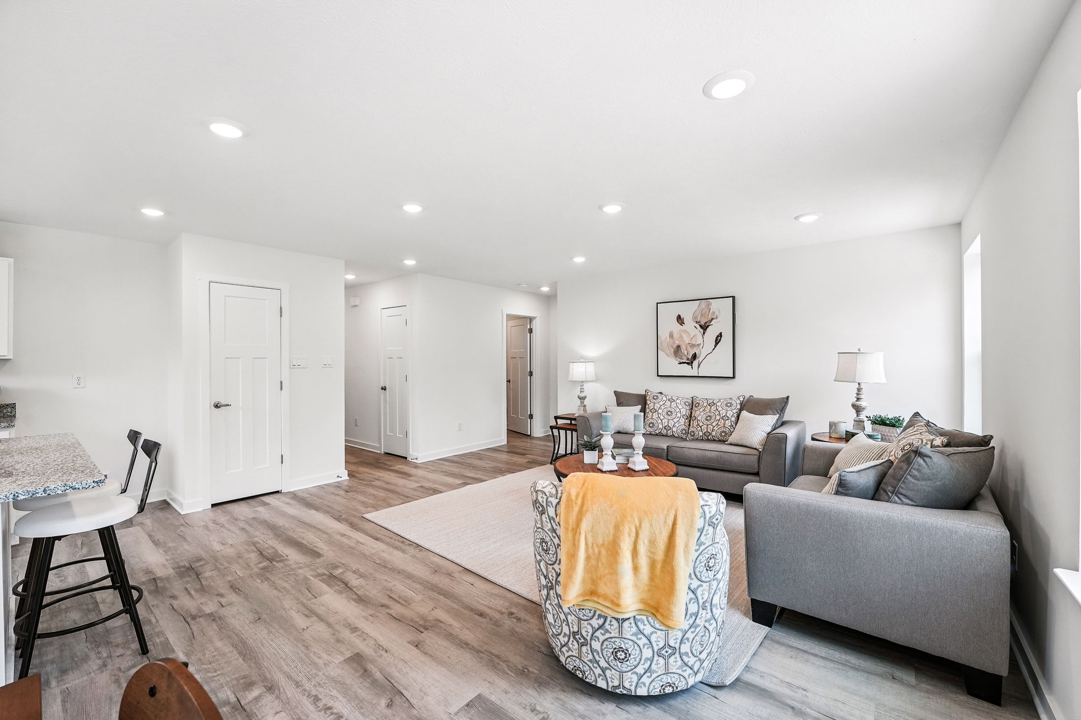 Modern living room with gray sofas, wooden flooring, and bright white walls, featuring stylish decor and recessed lighting.