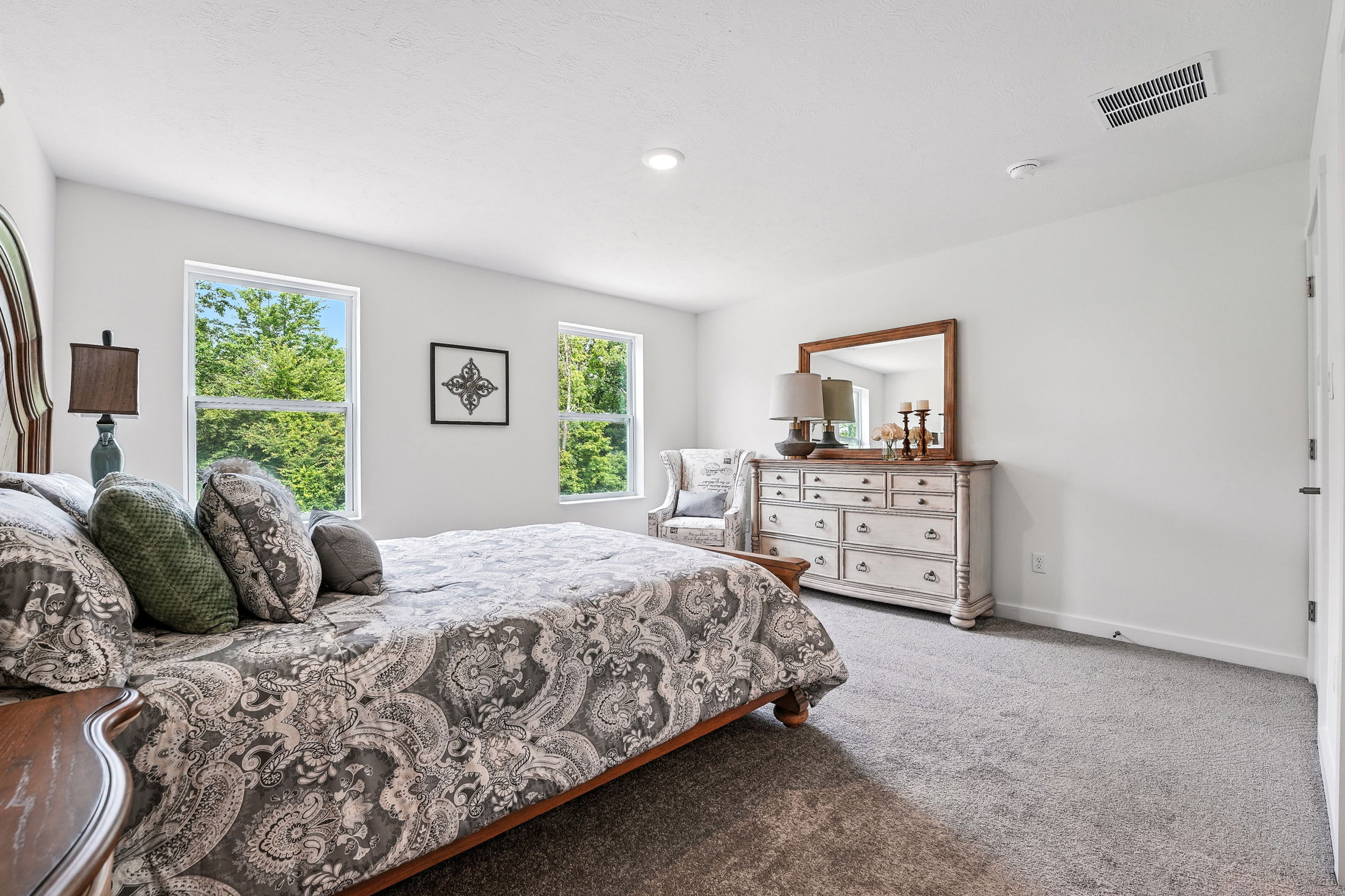 A cozy bedroom with a patterned bedspread, two windows, and a wooden dresser with a large mirror and decorative items.