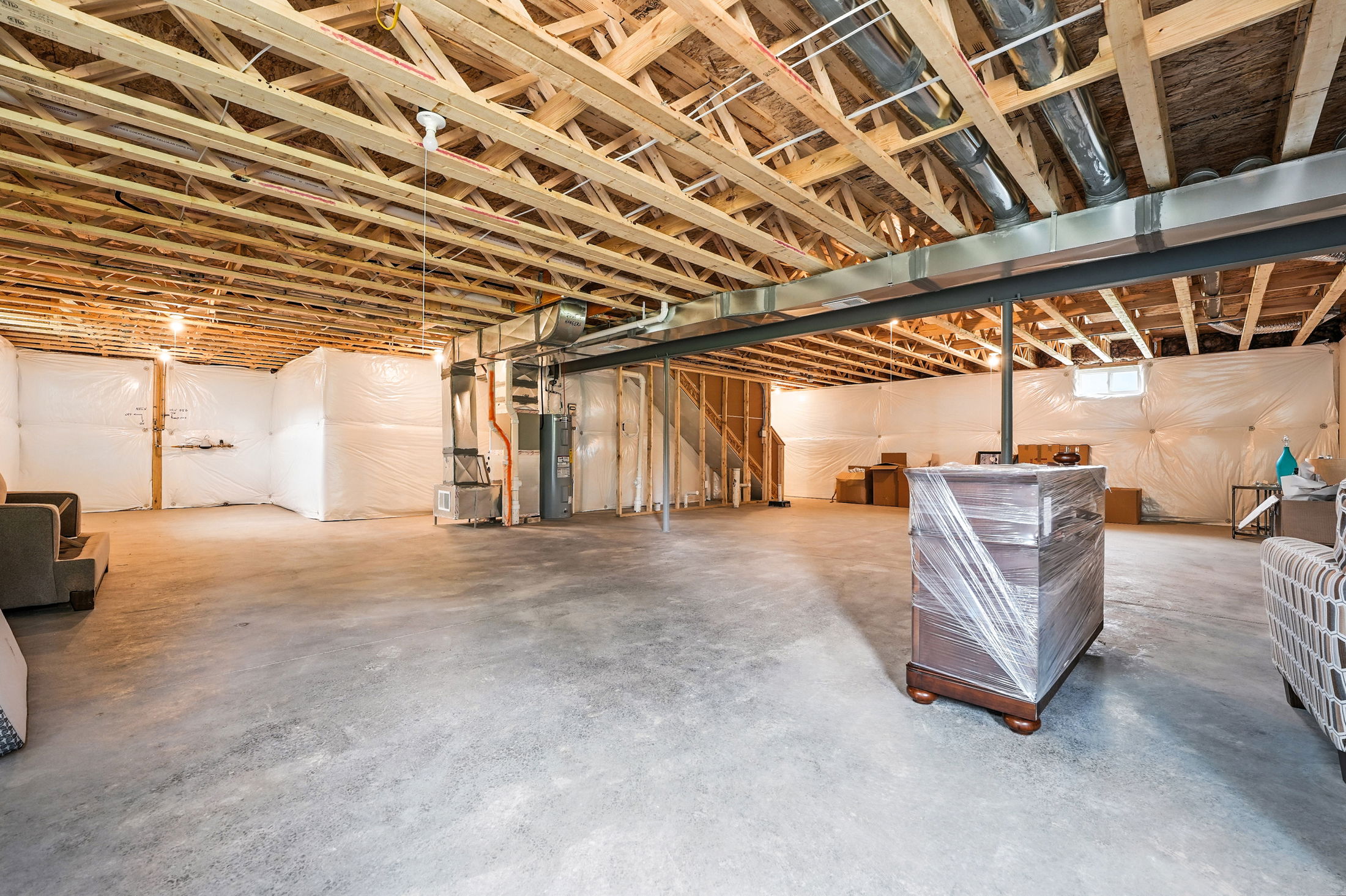 Unfinished basement with exposed wooden ceiling beams, concrete floor, and wrapped furniture.