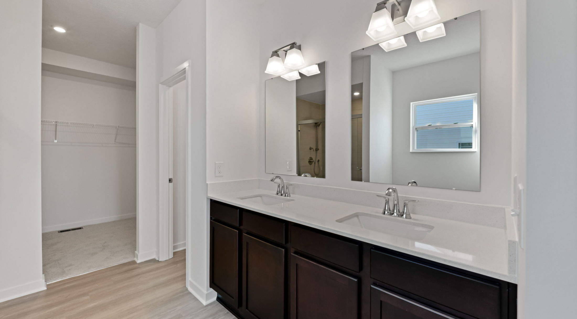 Modern bathroom interior featuring a double vanity with dark wood cabinets, white countertops, dual mirrors, and chrome faucets, adjacent to a walk-in closet.