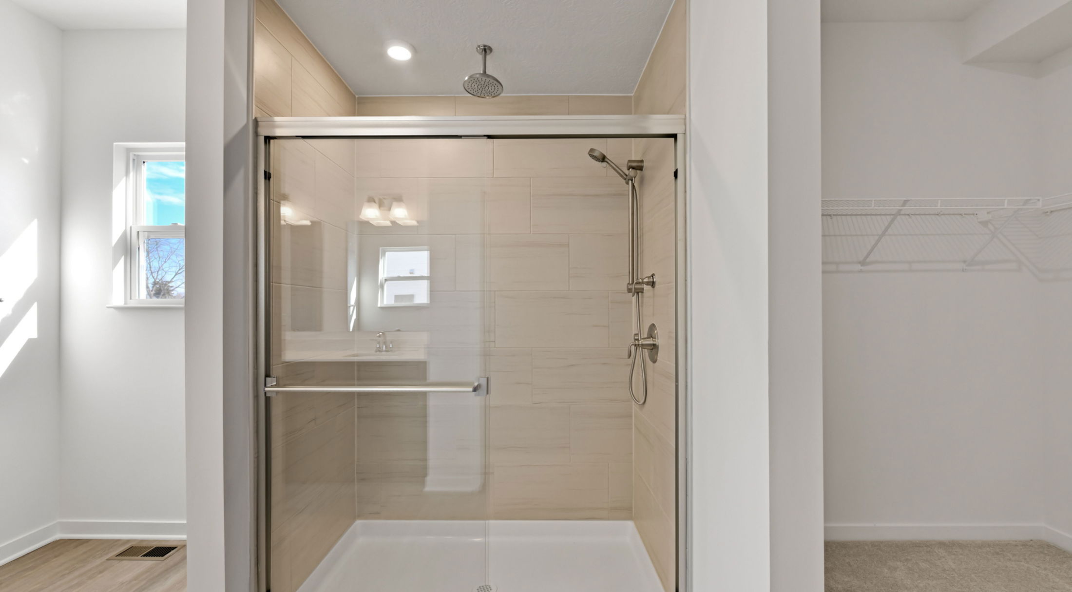 Modern bathroom with a glass-enclosed shower and beige tile walls, featuring dual showerheads and ample natural light.