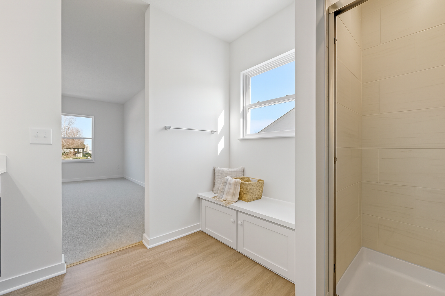 Bright and modern bathroom with wooden flooring, a cozy sitting area with cushions and a basket, adjacent to a walk-in shower and carpeted room.