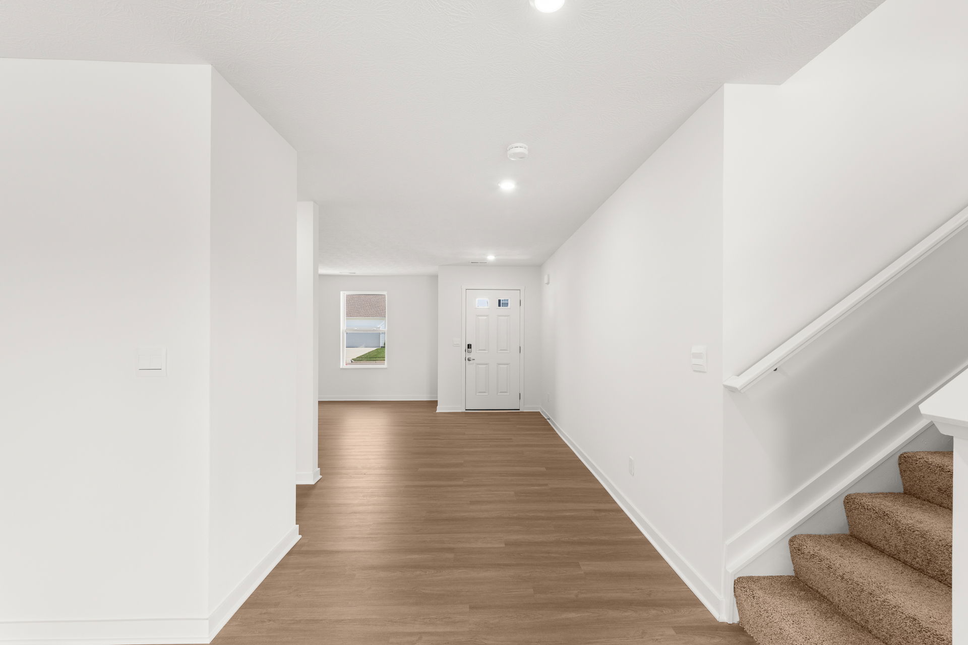 Modern, empty entryway of a bright interior home with white walls, wooden floors, and a carpeted staircase.