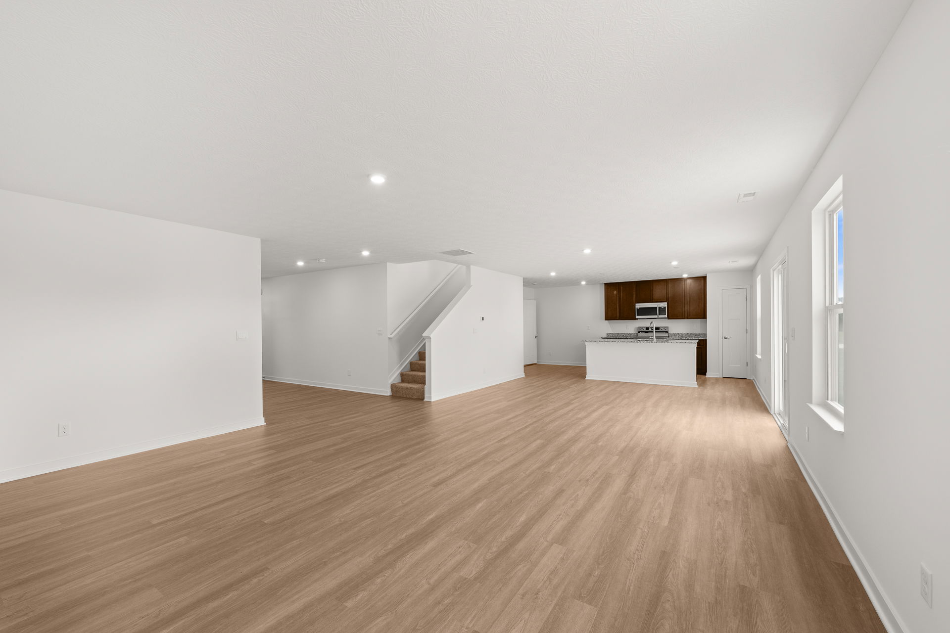 Modern open-concept living room with hardwood floors, a staircase, and an adjacent kitchen featuring dark wood cabinetry and stainless steel appliances.