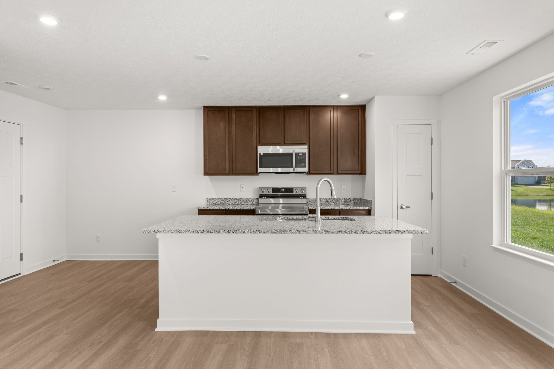 Modern kitchen with granite countertops, brown cabinets, stainless steel appliances, and a view of the outdoors through a large window.