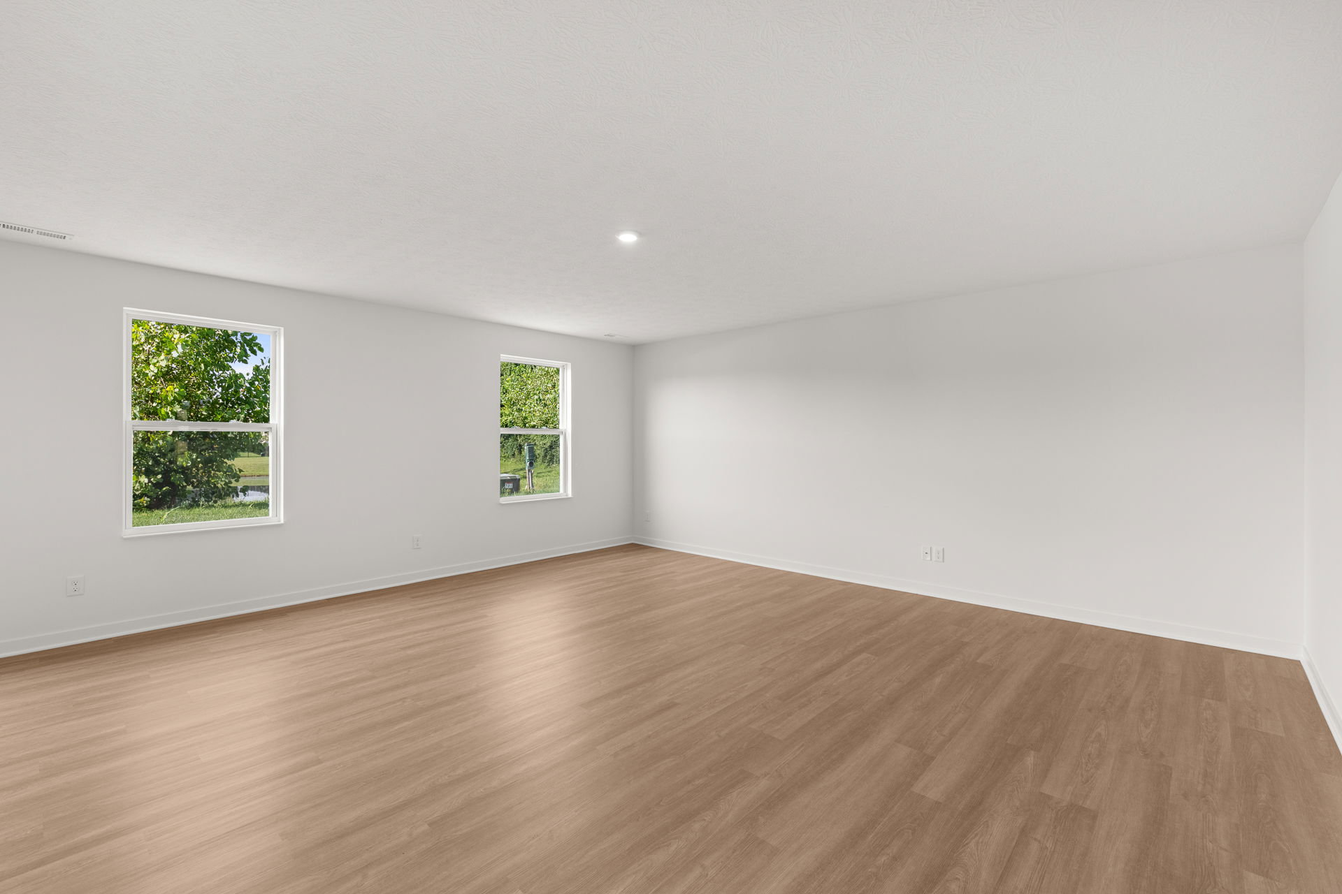 Spacious empty room with white walls, wood flooring, and two windows overlooking green trees.