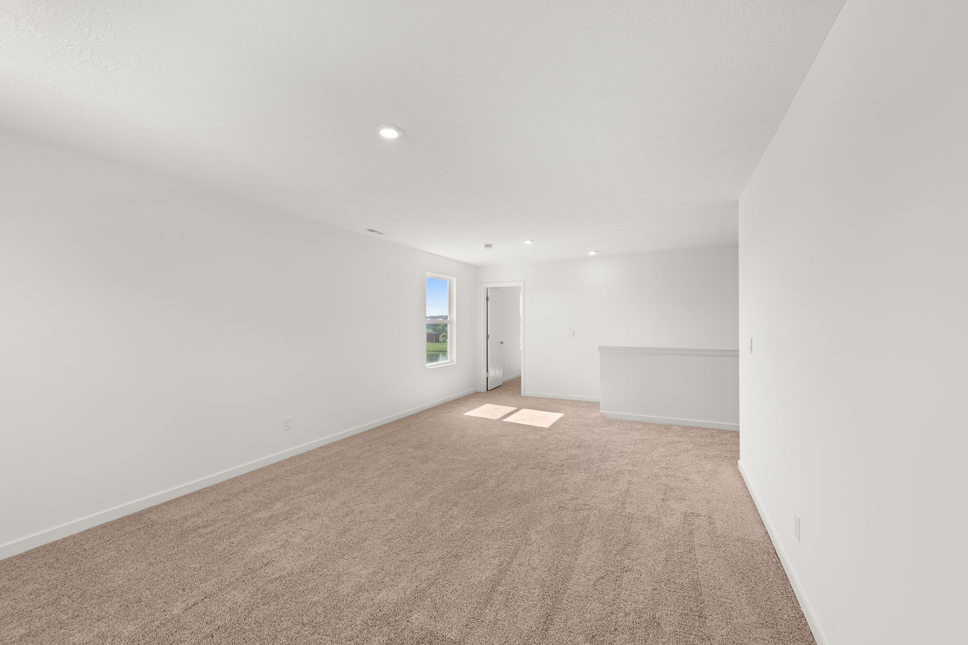 Spacious, empty room with beige carpet, white walls, recessed lighting, and a window providing natural light.