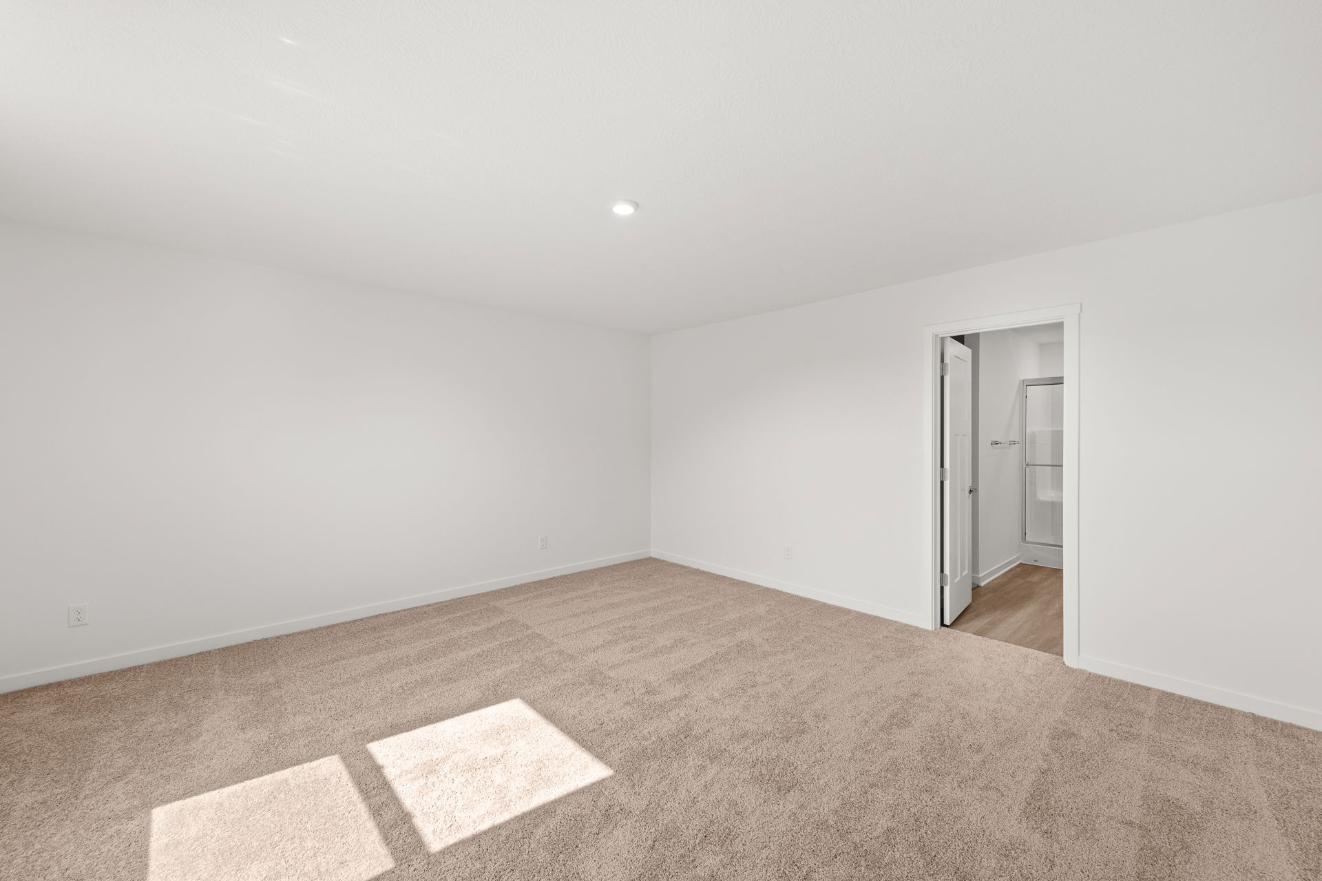 Spacious empty room with beige carpet, white walls, and an open door leading to a bathroom.