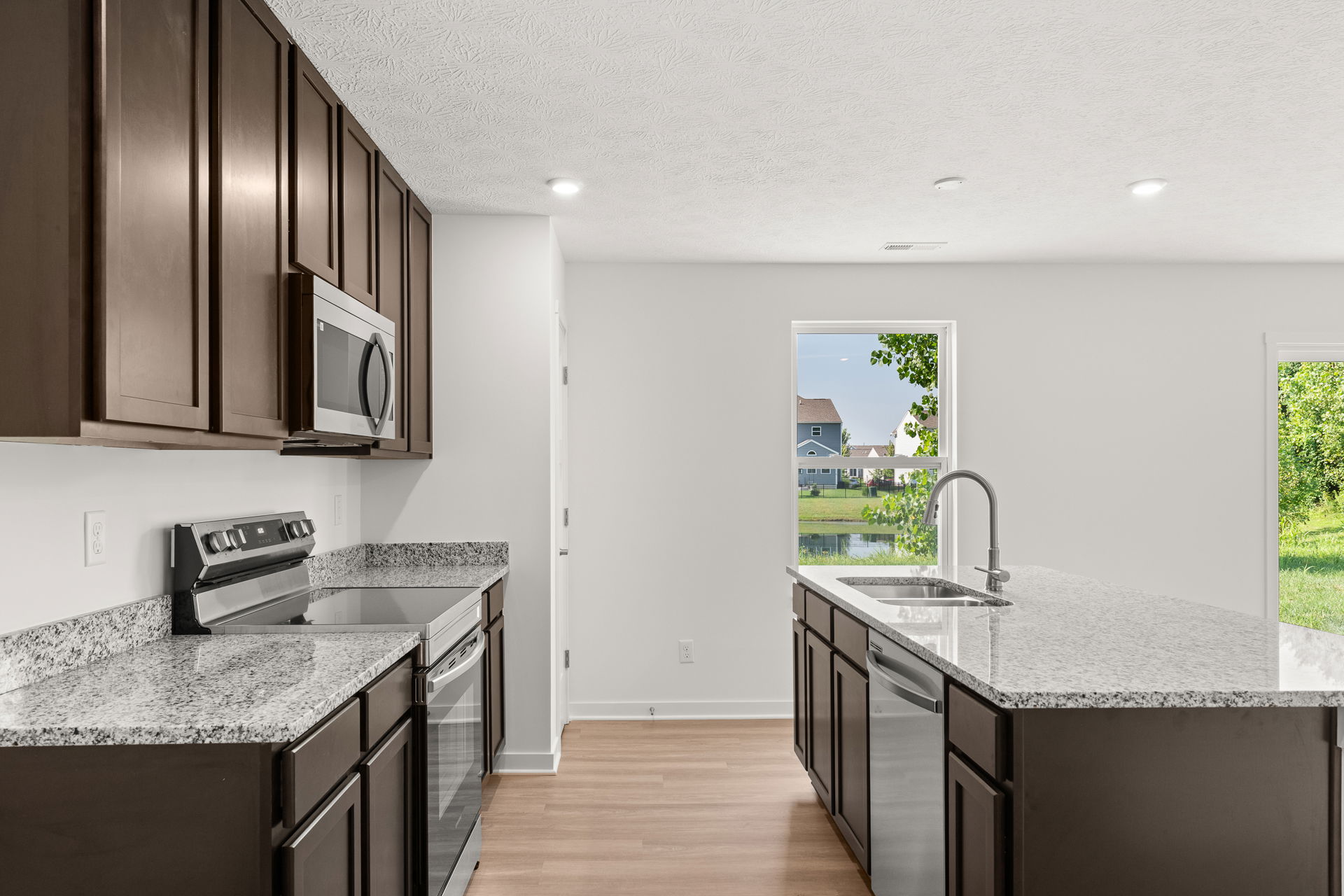 Modern kitchen interior with dark wood cabinets, granite countertops, and stainless steel appliances overlooking a scenic backyard view through large windows.
