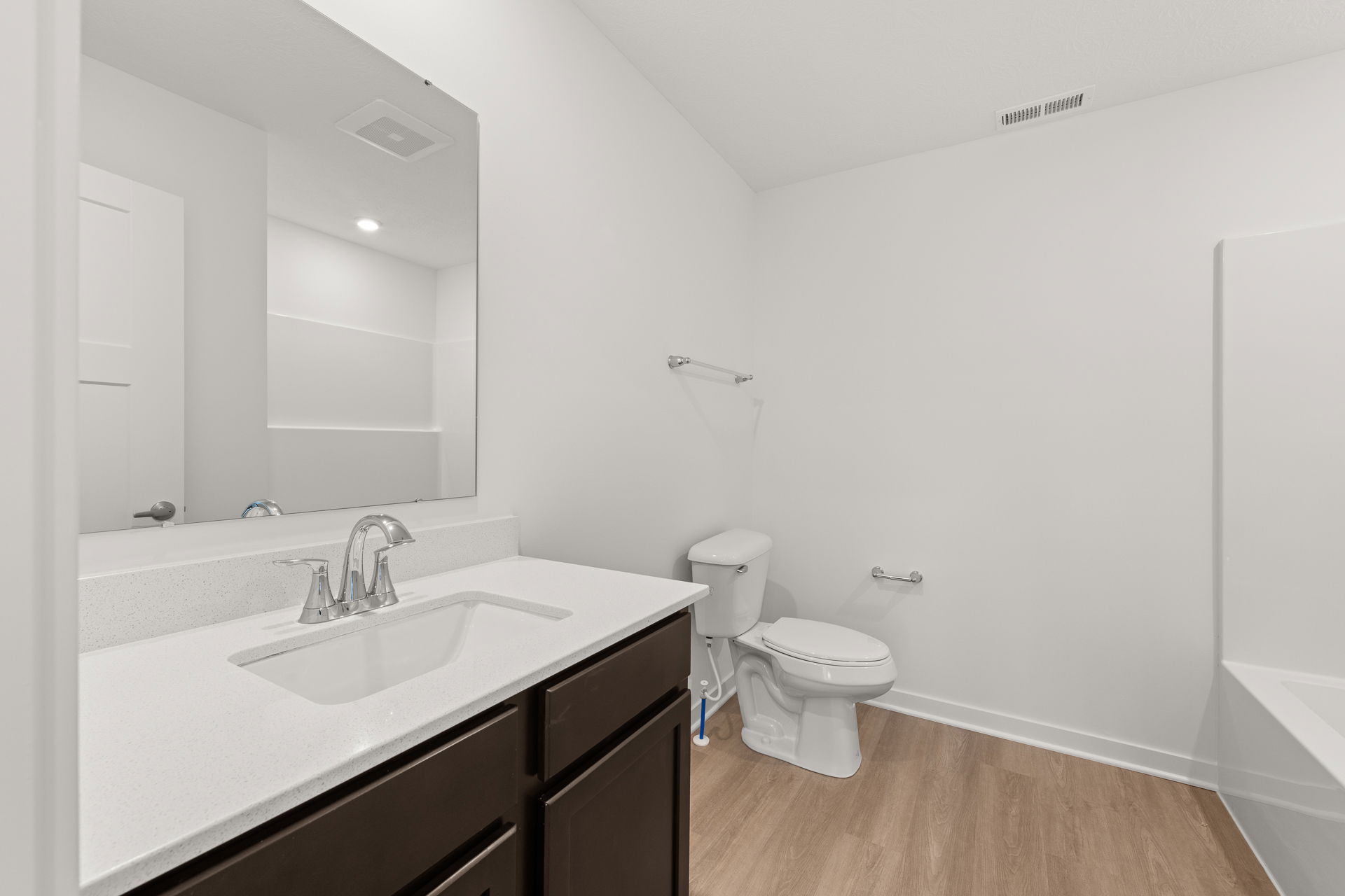 A modern bathroom featuring a white countertop, dark wood cabinets, a mounted mirror, and a clean, spacious design with light wood flooring.