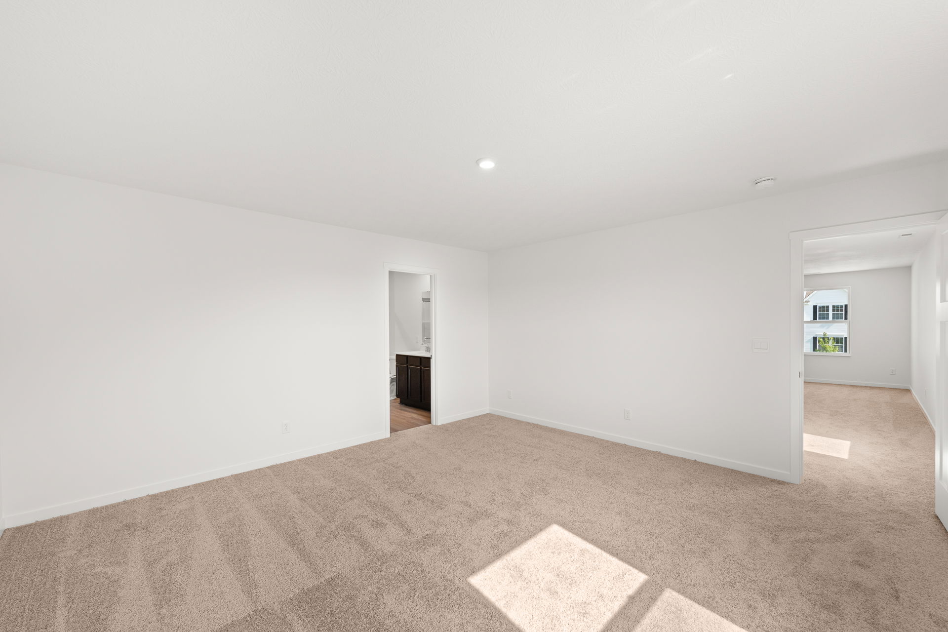 Spacious, empty room with beige carpeting and white walls, showcasing natural light and an open doorway to an adjacent room.