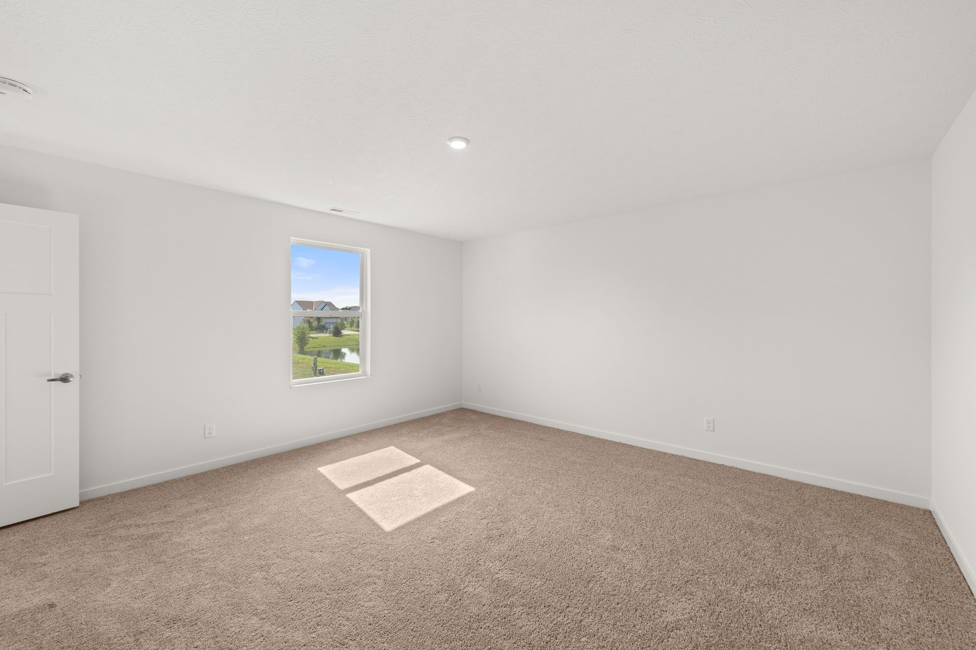 Spacious and bright empty bedroom with a window overlooking green scenery and soft beige carpet.