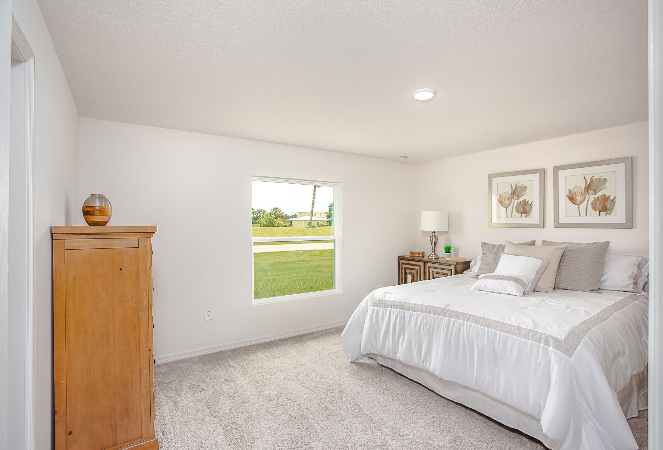 Bright and cozy bedroom with a large bed, wooden furniture, floral artwork, and a window overlooking a green lawn.