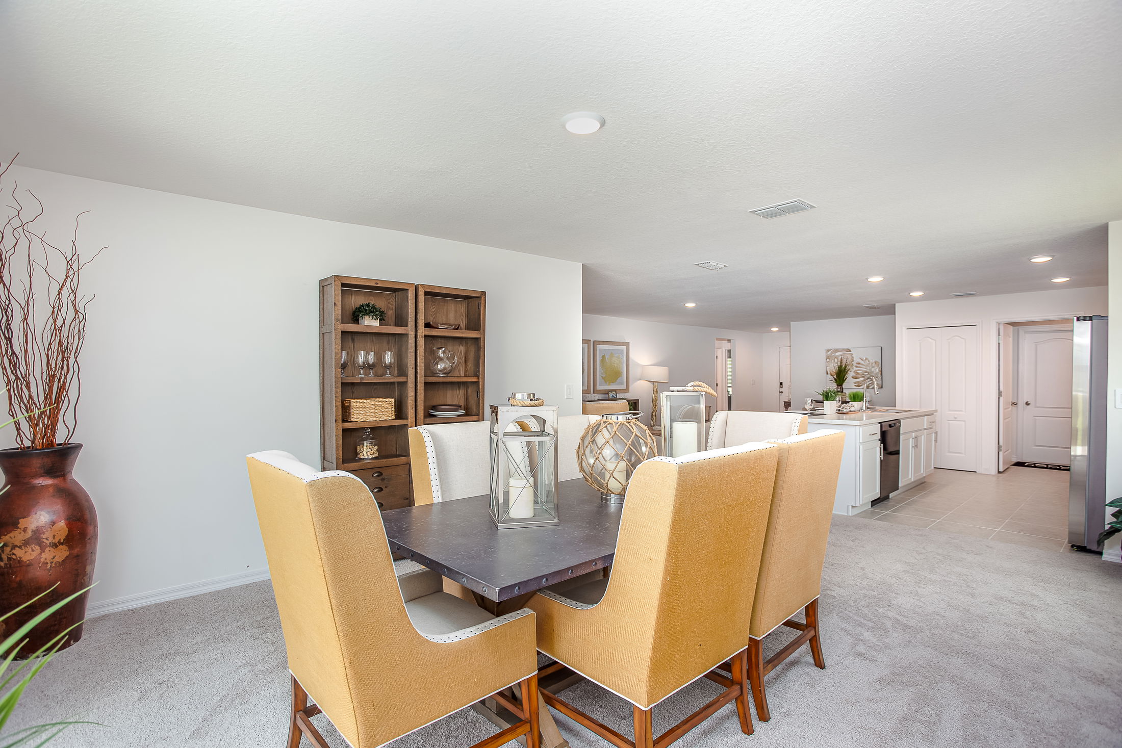 Modern dining room with beige upholstered chairs, a dark table, decorative lanterns, and an open view to the kitchen with white cabinets.