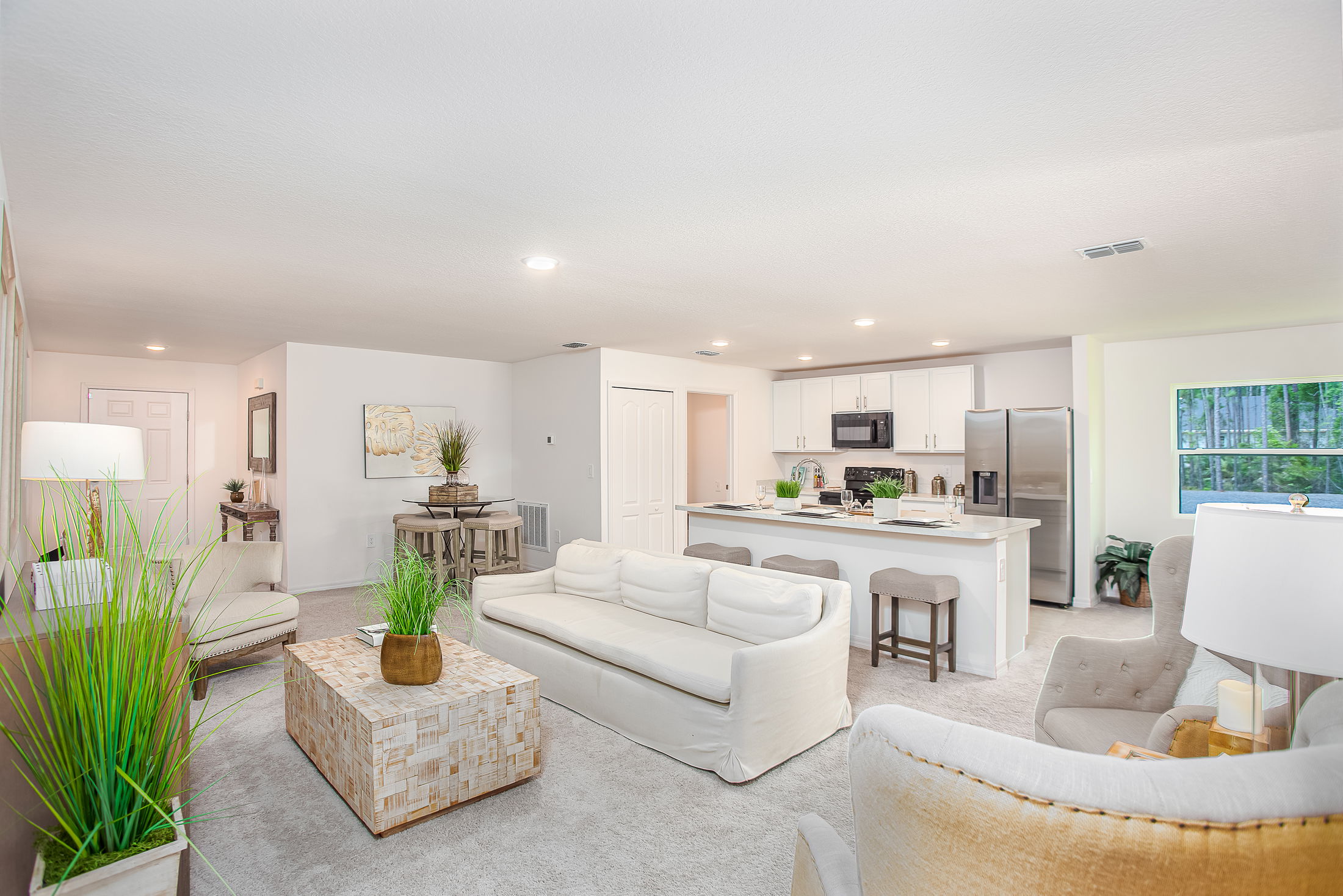 A modern open-concept living room and kitchen with neutral tones, white sofa, lush plants, and sleek appliances.