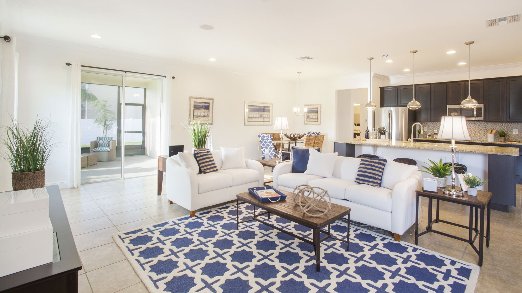 Bright and spacious open-concept living room with modern kitchen, featuring white sofas, blue geometric rug, and large sliding doors leading to a patio.