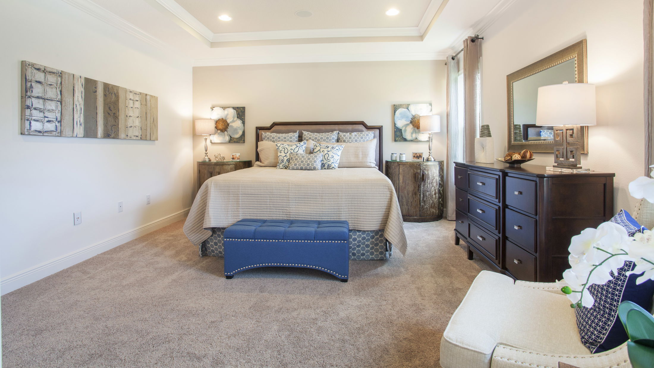 Elegant master bedroom featuring a cozy king-sized bed with patterned pillows, wall art, dark wood furniture, and a stylish blue ottoman.