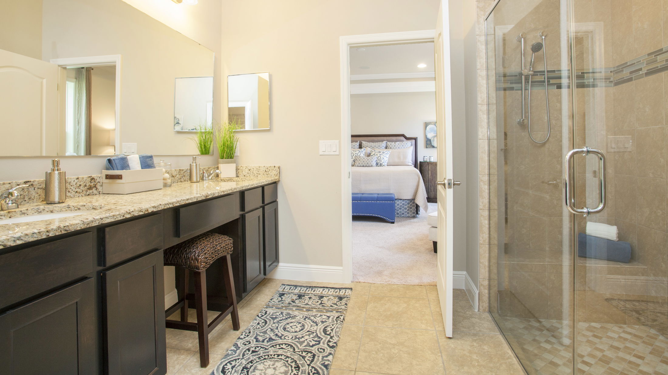 Modern bathroom with a glass-enclosed shower, granite countertop double sink, and doorway leading to a cozy, decorated bedroom.