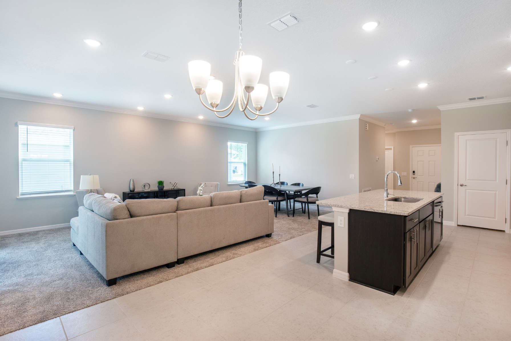 Spacious modern open-concept living room with a cozy beige sectional, elegant dining area, and sleek kitchen island under a stylish chandelier.