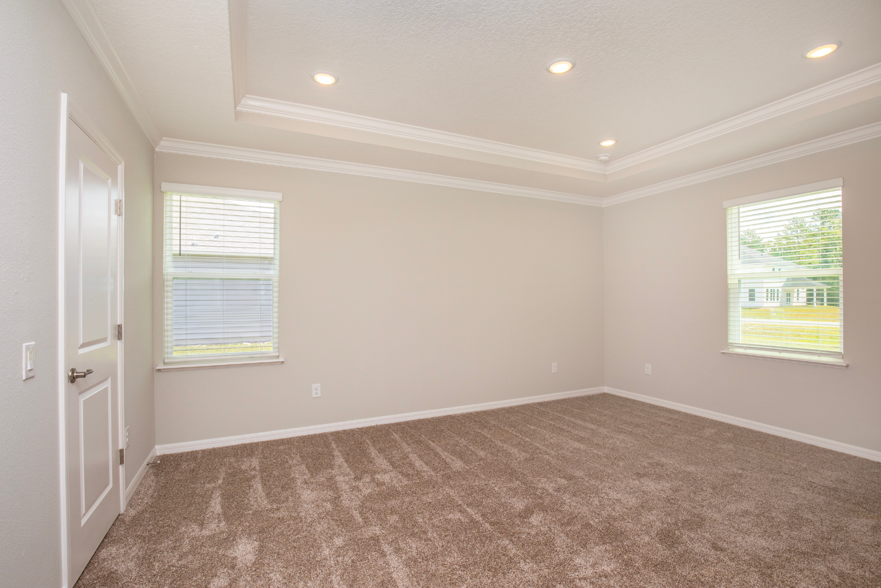 Empty beige room with carpet flooring, white trim, and recessed lighting, featuring two windows with blinds.