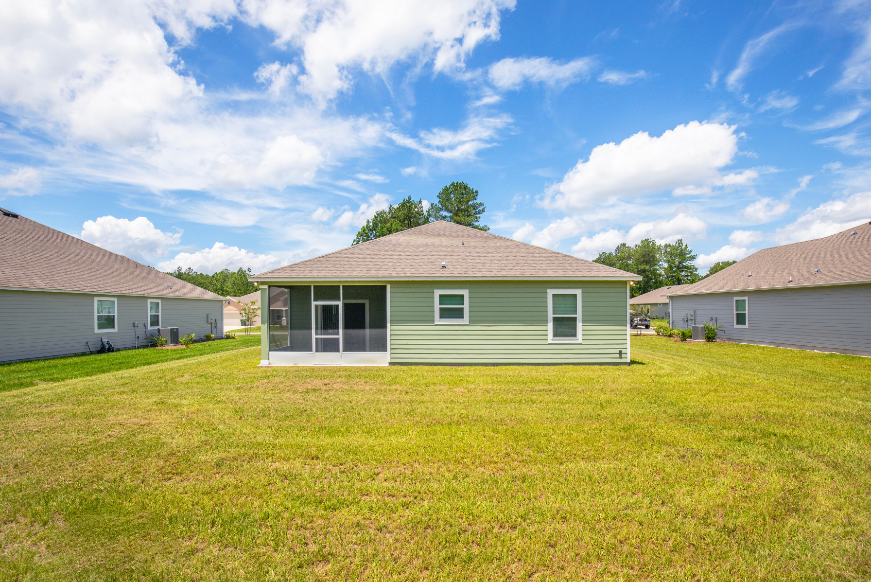 Rear view of a green suburban house with a screened patio and spacious lawn under a blue sky with clouds.