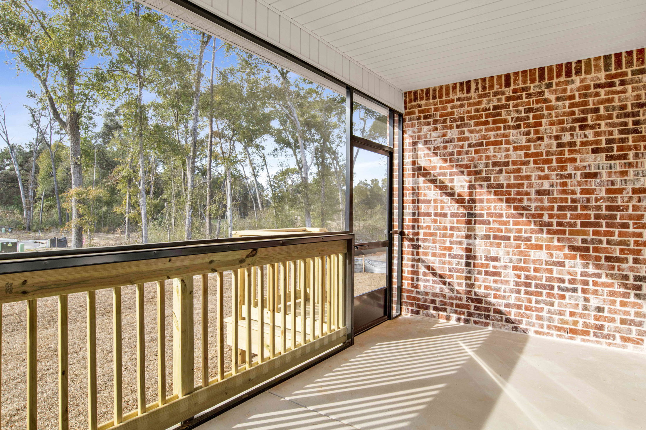 Screened-in porch with brick wall and wooden railing overlooking a wooded area in sunlight.