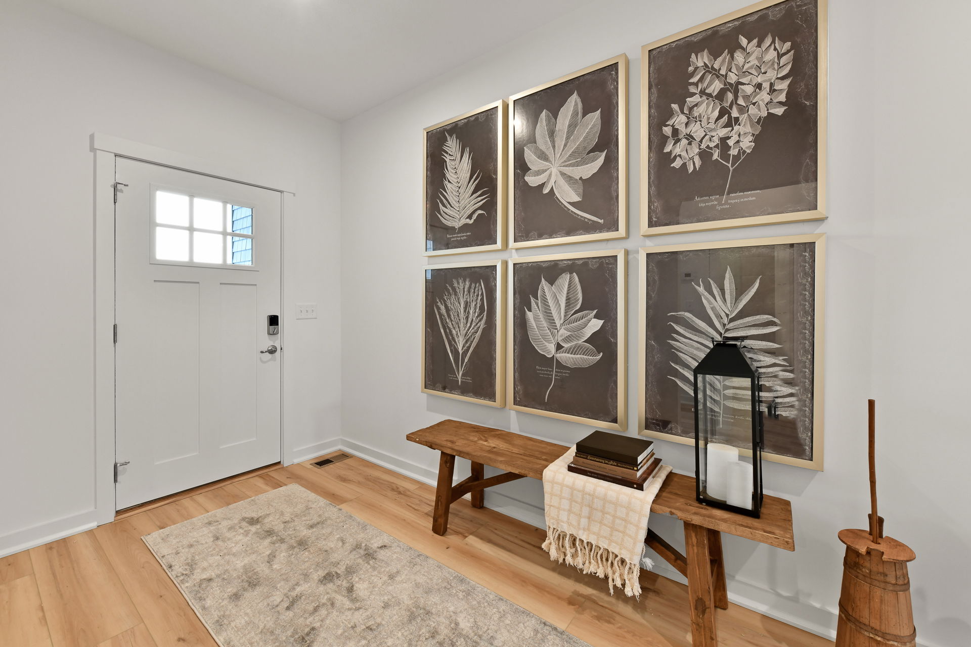 Modern entryway with framed botanical art, a wooden bench, books, lantern, rug, and rustic churn for a stylish welcome.