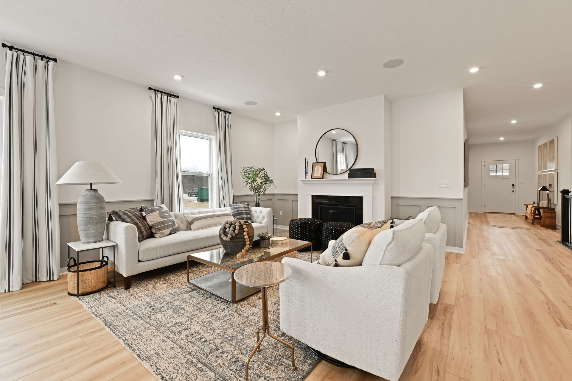 A modern, elegantly decorated living room with a light color scheme, featuring white sofas, a wooden coffee table, a large area rug, and a fireplace with a round mirror above it.