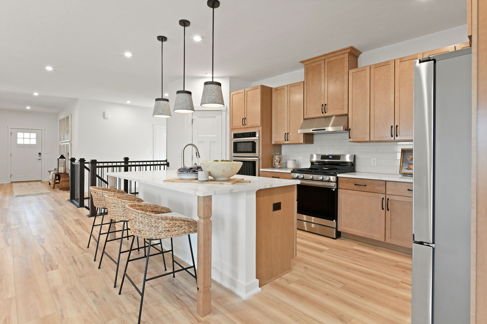 Modern kitchen with light wood cabinets, a white island, stainless steel appliances, pendant lighting, and woven barstools on hardwood flooring.