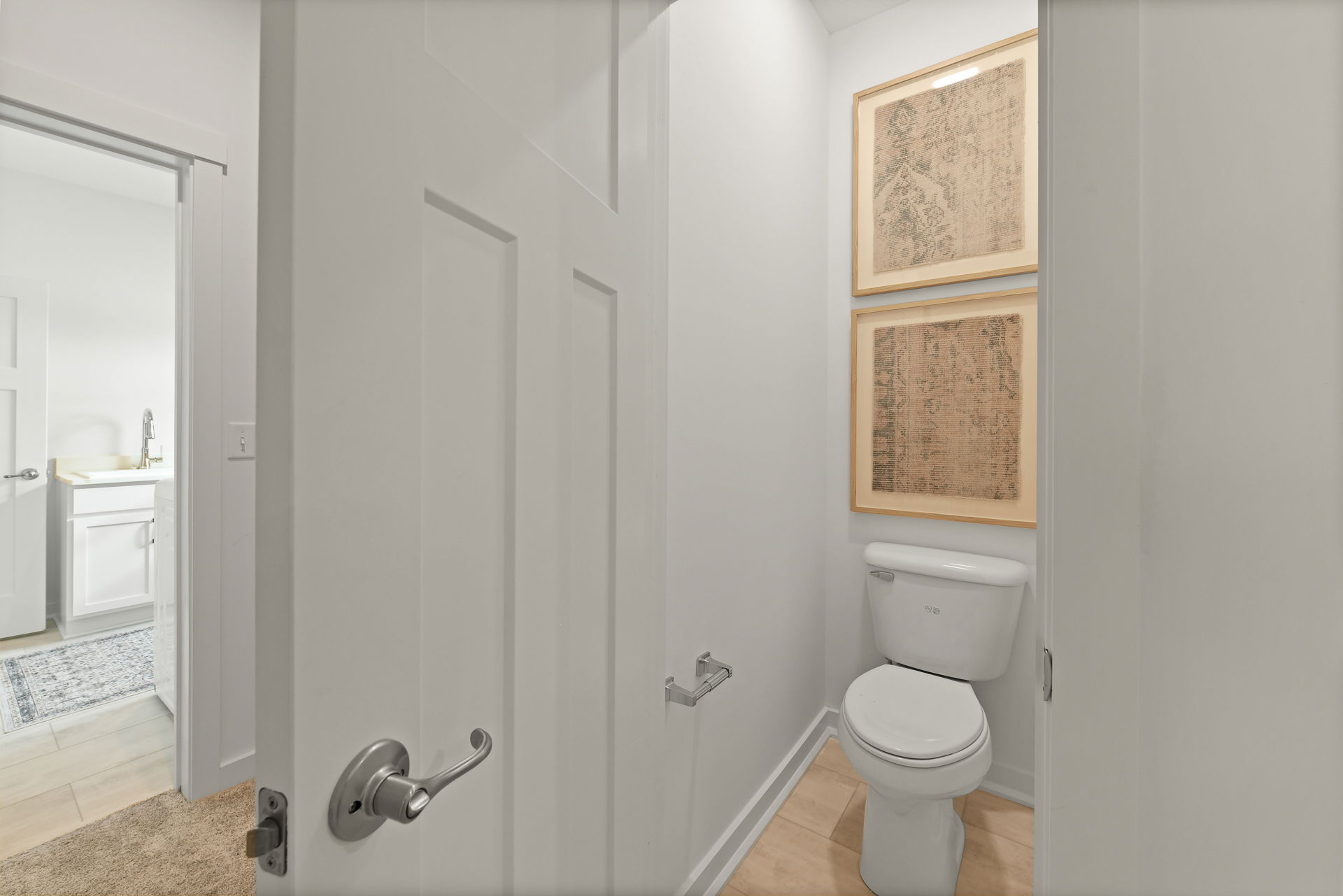 Small modern toilet room with white walls, framed wall art, and adjacent bathroom sink area.