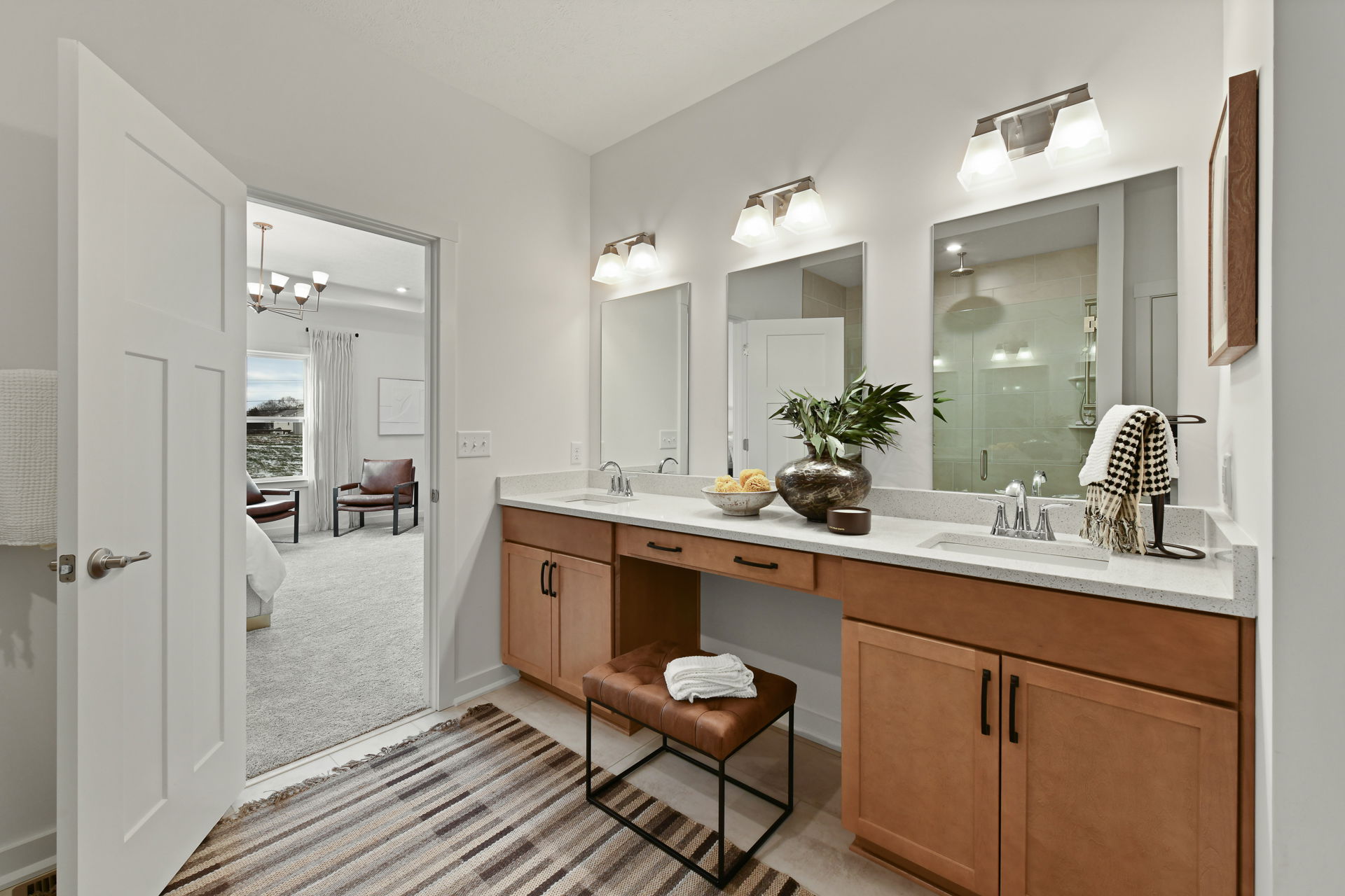Modern bathroom with twin sinks, wooden cabinetry, a decorative plant, and a view into an elegantly furnished bedroom.