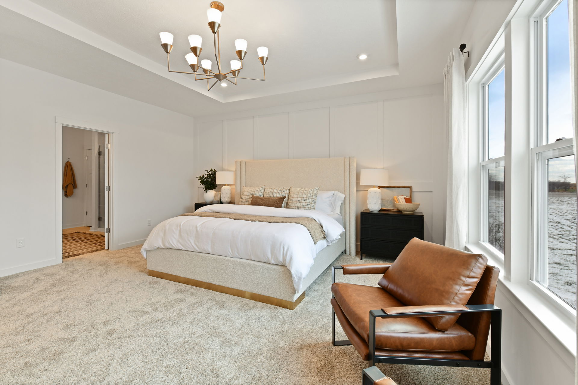 Modern bedroom with elegant chandelier, upholstered bed, leather chair, and large window with natural light.