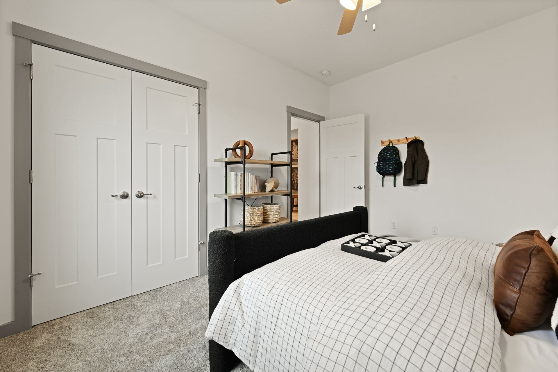 Modern bedroom with white walls, a black upholstered bed, patterned bedding, a ceiling fan, double closet doors, and minimalist decor elements.