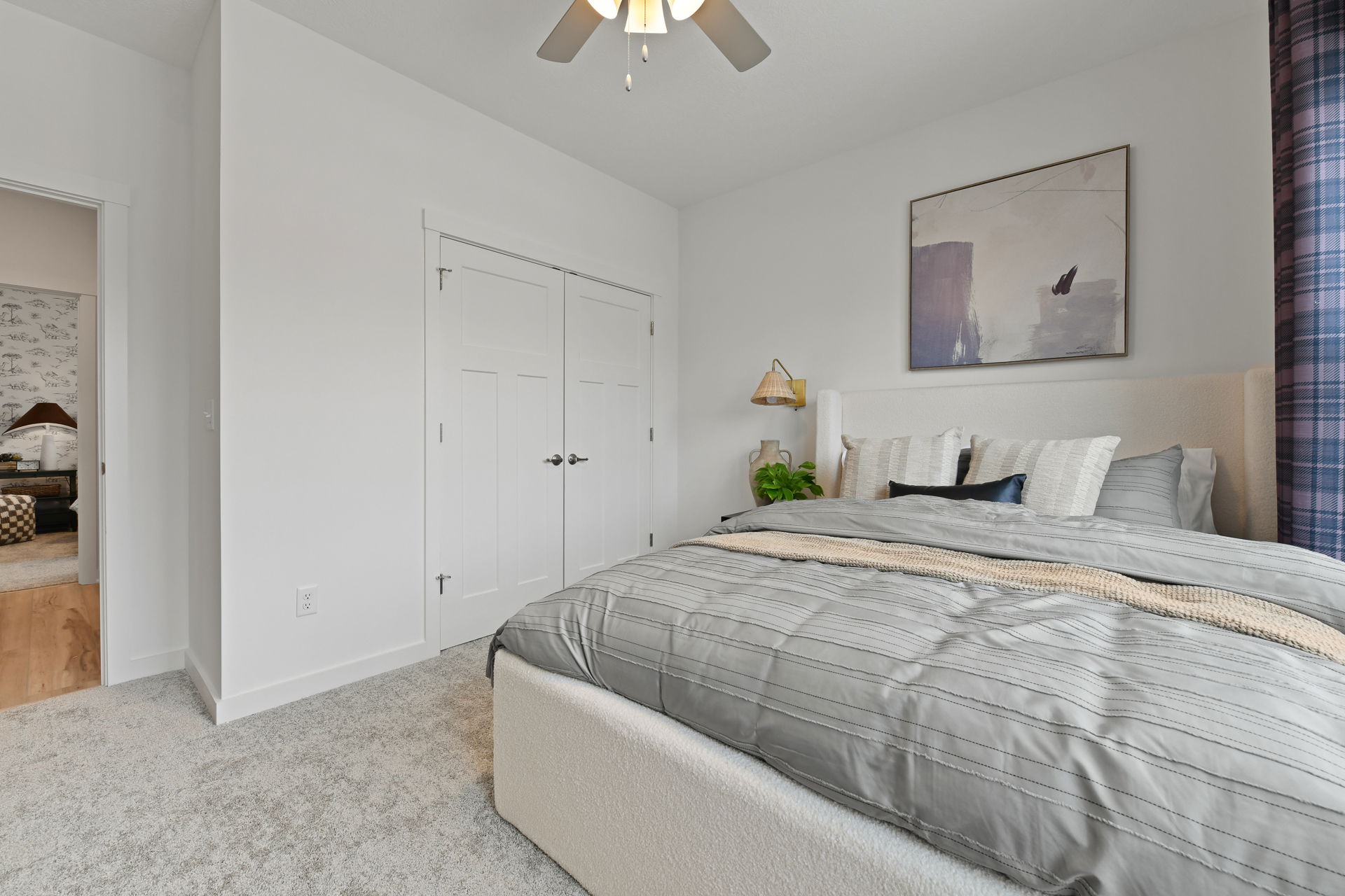 Modern bedroom with a cozy bed, striped gray bedding, abstract artwork, and a plant, featuring white walls and a ceiling fan.